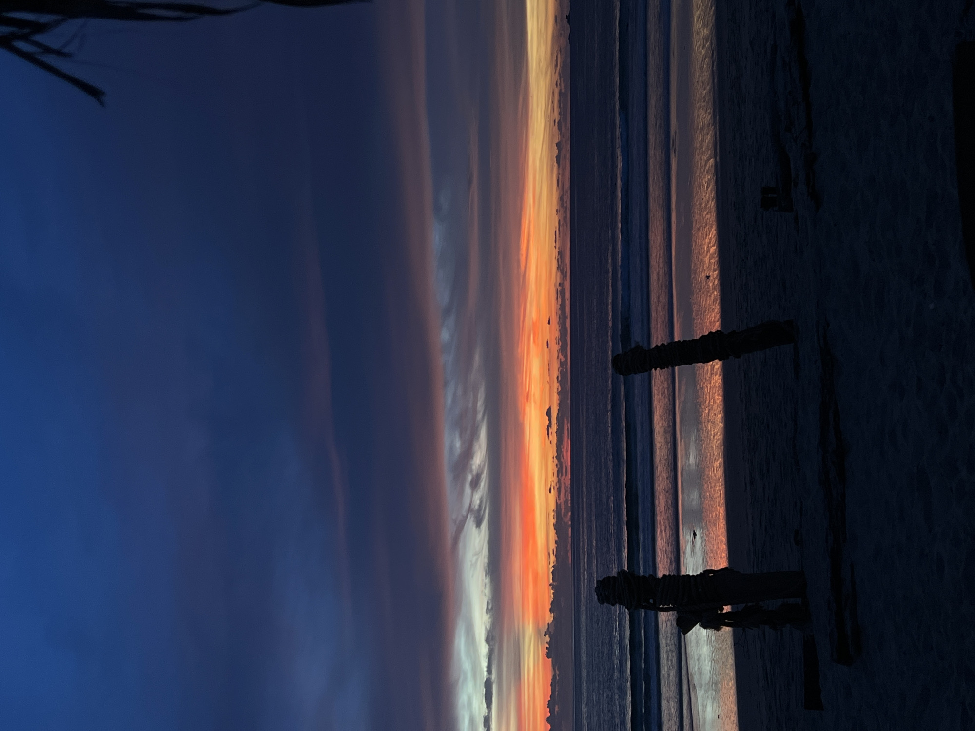 An outdoor, low-angle, vertical shot captures the twilight sky over a beach. The sky is a deep blue at the top, fading into vibrant hues of orange and pink closer to the horizon, indicating sunset. Clouds are visible in bands across the sky, some illuminated by the setting sun. The ocean is dark blue, with gentle waves creating white streaks on the surface. The wet sand on the beach reflects the colors of the sky. In the foreground, silhouetted against the light, are two weathered wooden posts wrapped in rope, resembling primitive structures or remnants of a pier. The ground is covered in dark sand. A dark, out-of-focus object, possibly foliage or a structure, is present in the upper right corner.