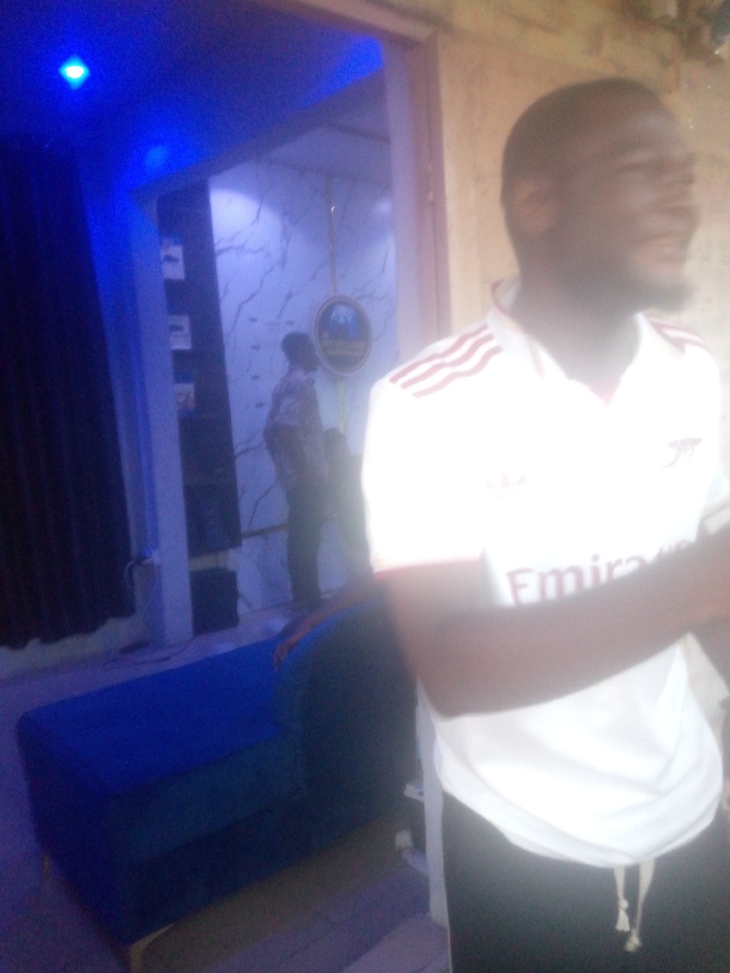 The image captures a dimly lit indoor scene with blue lighting. In the foreground, a man wearing a white Arsenal jersey with red stripes and the "Emirates" logo is visible, facing away from the camera and to the right. He has short dark hair and a beard. Behind him, another person is standing in a doorway or passage, wearing a patterned shirt and dark trousers. To the left, a dark blue sofa or couch is situated. The background reveals a wall with a marble-like pattern and some shelves. The overall impression is that of an entertainment venue or a private residence with a recreational area, possibly located in Jalingo, Nigeria.