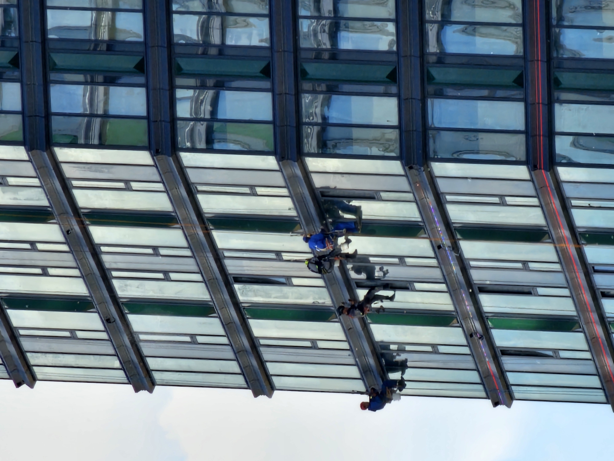 This is a high-angle, rotated photograph capturing a segment of a modern glass and steel skyscraper facade in Moscow, Russia. The image is oriented such that the building appears to extend horizontally, with the sky filling the lower left portion of the frame.
Several individuals, identified as rope access technicians or window cleaners, are suspended on ropes against the building's exterior. They are actively engaged in descending the facade, wearing blue work uniforms, safety harnesses, and helmets, indicative of their specialized task. One person in a blue jacket is prominently featured in the upper middle, while two others are further down and to the right, with their reflections visible in the glass panels.
The building exhibits a complex architectural design, featuring alternating horizontal bands of dark, reflective glass and lighter, possibly frosted or opaque, panels. Some glass sections appear tinted green, and reflections of the sky and surrounding environment distort the glass surfaces. Diagonal structural elements divide these panels, creating a dynamic visual pattern. Bright red lines run vertically along some of the darker structural beams on the right side of the building.
The photograph is taken in bright daylight under clear weather conditions, as evidenced by the clear sky visible. The perspective is from below, looking up at the towering structure and the workers, giving a sense of scale and the height at which they are operating. There is no visible text in the image.