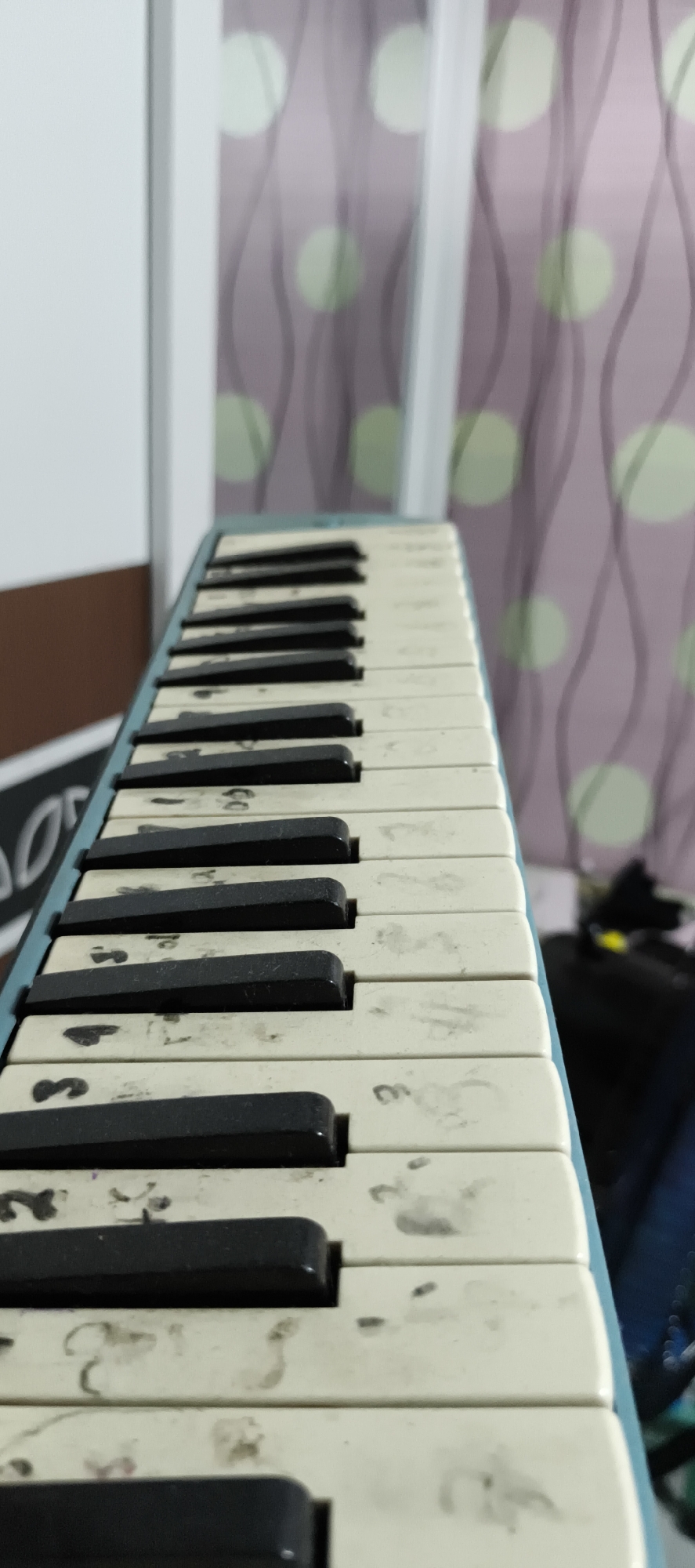The image shows a close-up, side-view of a melodica's keyboard. The keys are white with black button-like separators. Some of the white keys have smudges and what appear to be handwritten numbers or musical notation, such as "3" and "2". The melodica is blue on the left side. In the background, there is a light-colored wall on the left and a curtain with a pattern of wavy lines and circles on the right. The lighting suggests an indoor setting. There is no visible indication of the specific location within Muaro Jambi, Indonesia.