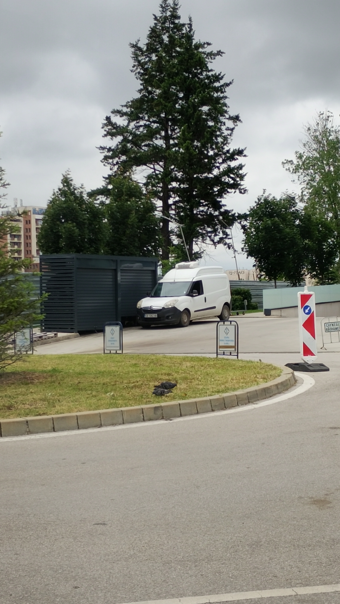 A white panel van with a refrigerated compartment on top is parked on a paved surface next to a dark gray, slatted structure. Two dark-colored signs on metal stands are placed in front of the van and structure. The van has a license plate that reads "CB 1082 AR". In the background, there are trees, a cloudy sky, and a building with multiple floors. To the right of the van, a red and white striped traffic cone with a blue arrow sign is positioned. The foreground shows a paved road curving to the right, bordered by a grass median with several pigeons.