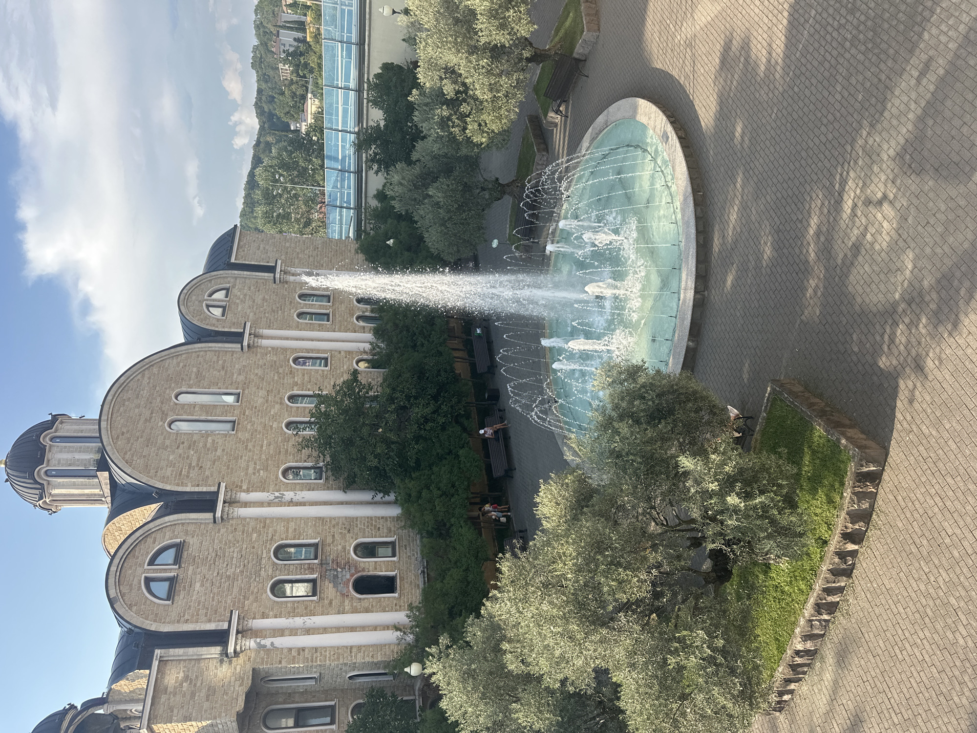 This is an eye-level, medium shot of a public square in Sochi, Russia, featuring a large, multi-tiered fountain at its center. The fountain's water is illuminated and appears a vibrant turquoise color, with jets spraying high into the air. The square is paved with gray interlocking bricks, and stone planters containing grass and olive trees are strategically placed around the fountain.
Dominating the left side of the frame is a large, ornate building with a light-colored stone facade. The building has a multi-domed, dark roof, arched windows of various sizes, and classical-style white columns supporting the upper sections. The architecture suggests a religious or historical significance.
In the background, lush green trees and rolling hills are visible, with some buildings scattered on the slopes. A modern blue-glass structure, possibly a bridge or walkway, is present in the upper right portion of the background.
Several people are present in the scene. On the left side, near the building, a group of three individuals, likely adults, are standing together. Further to the right, several people are seated on dark-colored park benches placed along the perimeter of the square. One person is sitting alone on a bench near the fountain, wearing a white cap. The lighting suggests it is daytime, with the sun casting shadows across the brickwork. The overall atmosphere appears tranquil and inviting.