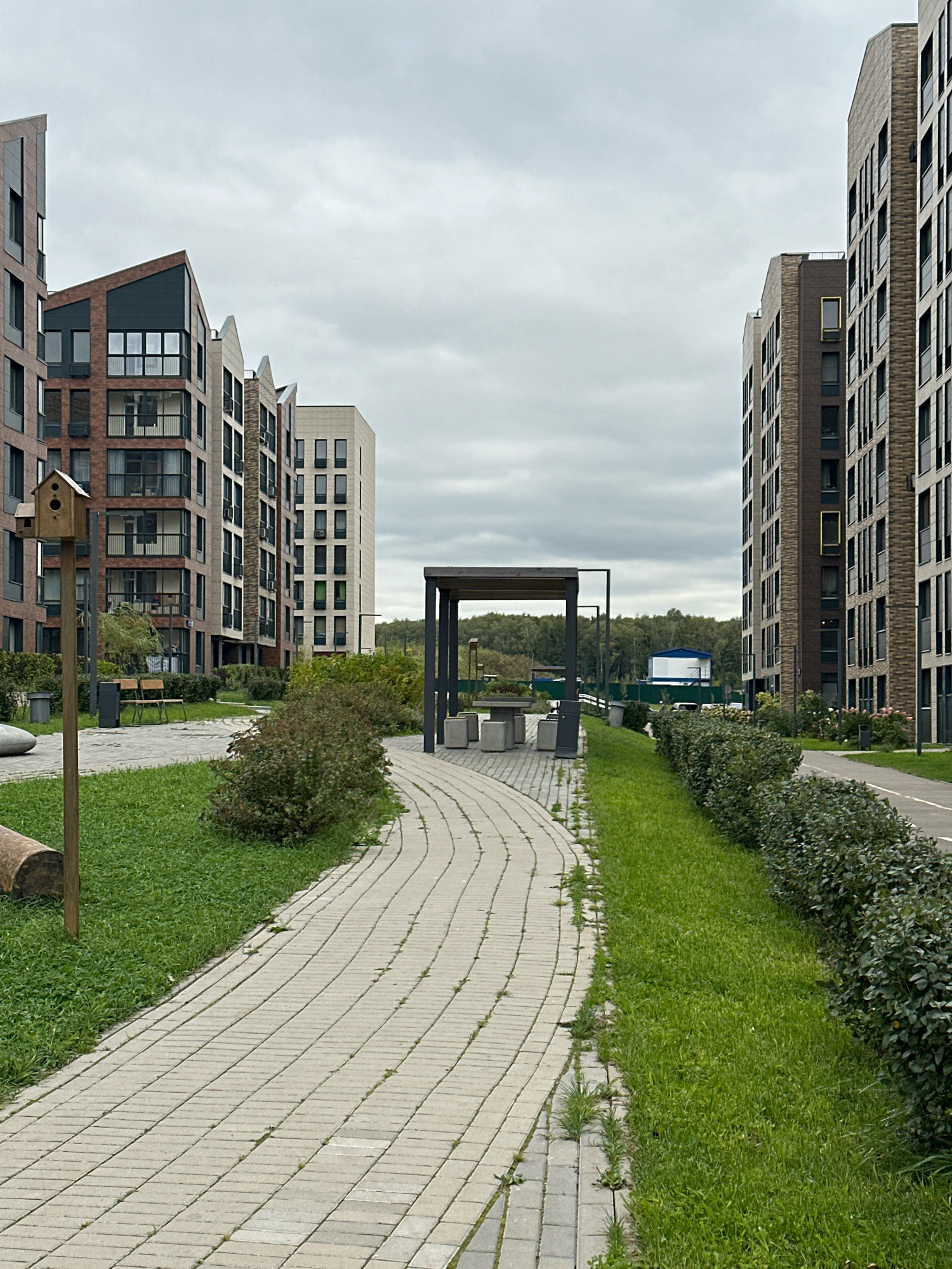 The image depicts a modern residential complex in the city of Vidnoye, Russia, under an overcast sky. In the foreground, a curved pedestrian path made of light-colored paving stones winds through a grassy area with bushes. To the left of the path, a birdhouse on a wooden pole stands near a large, light-colored rounded object, possibly a decorative stone. Further back, two benches are visible amidst greenery.
Dominating the scene are tall, multi-story apartment buildings with contemporary architecture. The buildings feature a mix of brick and light-colored facades, with numerous windows and balconies. The building on the left has a distinctive angled roofline. On the right, a similar tall building has a darker brick facade.
In the mid-ground, a covered seating area with a table and benches is situated along the path. Beyond the buildings, a line of trees forms a distant natural border, with a white and blue structure visible in the far distance. The overall environment suggests a well-maintained urban residential area. No people are present in the image.
