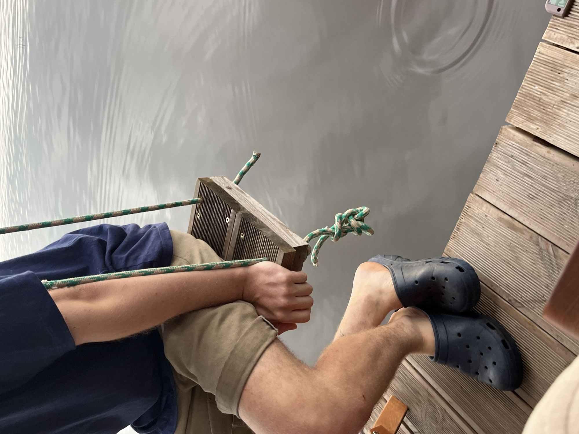 In Radchenko, Russia, a person sits on a wooden swing suspended over a body of water. The person wears a navy blue t-shirt and tan shorts, along with dark blue clogs. Their hands rest on their lap. The swing is made of a wooden plank and is attached to two green and white ropes, one of which is frayed at the end. The water's surface is calm with slight ripples. The scene appears to be a dock or pier.