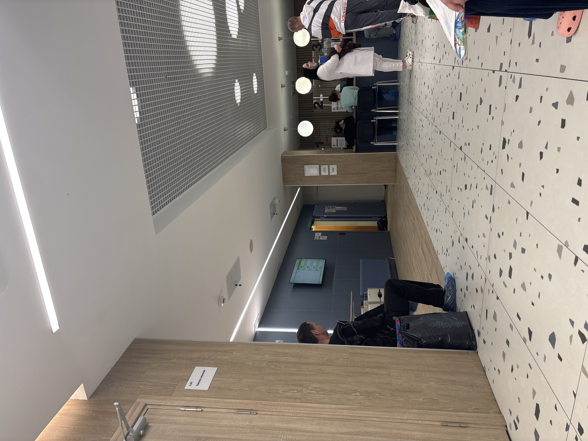 The image shows the interior of what appears to be a medical facility or clinic, potentially located in Saint Petersburg, Russia. In the foreground, a man is lying on the floor with a black plastic bag next to him. He is wearing dark clothing and blue shoe covers. To the right, the floor is tiled with a pattern of white and grey speckled tiles. Along the back wall, there is a blue wall with a digital display showing queue numbers. Further down the hallway, there are several people sitting in chairs. In the background, there are staff members behind a reception desk attending to patients. The ceiling has a grid-like structure with recessed lighting. A sign on the door on the left side reads "2.20" in Cyrillic. Overall, the scene depicts a waiting area, possibly in a public healthcare setting.