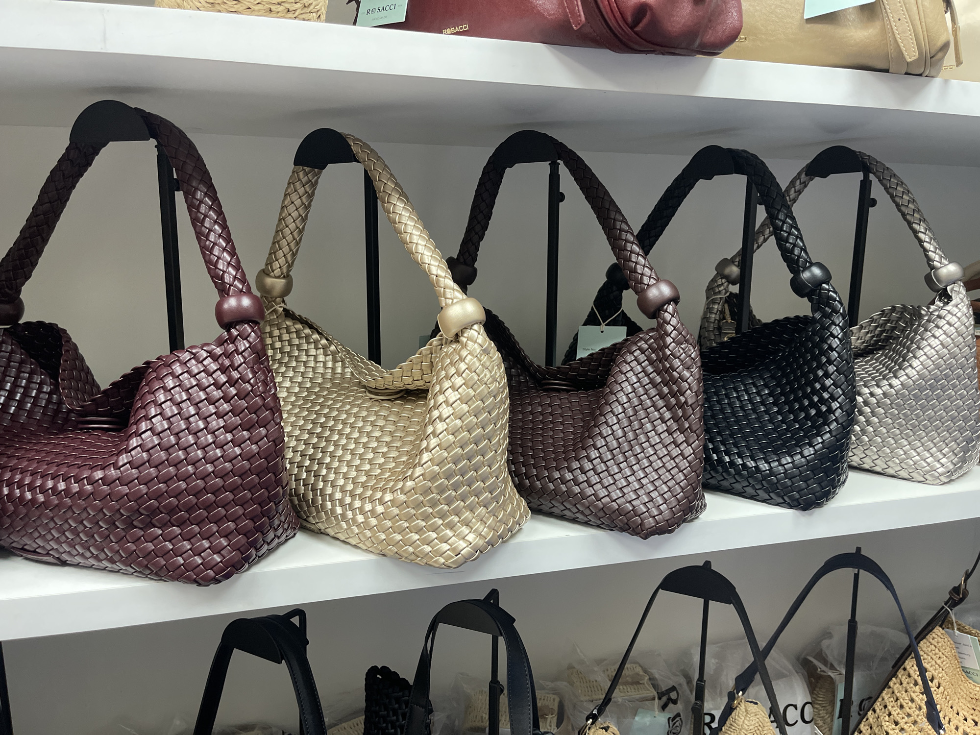 The image displays a collection of woven handbags on white shelves. The handbags are arranged in a row, with five distinct bags visible on the top shelf and several more partially visible on the bottom shelf. The handbags vary in color, including burgundy, gold, brown, black, and silver. Each bag features a braided handle and a woven texture. The background is simple and appears to be a retail environment. There are no people or animals present in the image. The lighting is even, suggesting an indoor setting. The image does not provide any information that would suggest the location is Moscow, Russia, or any other specific city.
