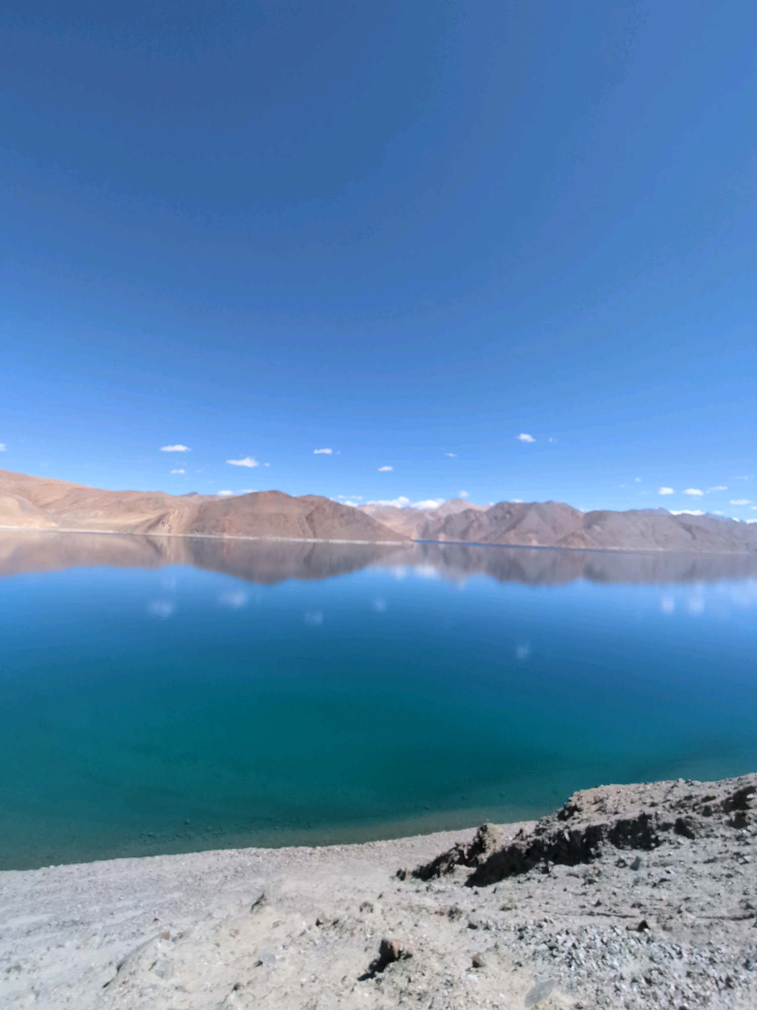 The image depicts a serene landscape in Lukung, India, featuring the stunning Pangong Tso lake. The upper half of the image is dominated by a vast expanse of clear blue sky, with a few wispy white clouds scattered across it. Below the sky, majestic mountains with rugged, brown and tan slopes stretch across the horizon. These mountains are beautifully reflected in the calm, turquoise waters of Pangong Tso, creating a mirror-like effect. The foreground of the image shows a rocky, barren shoreline with scattered stones and sand. The overall scene conveys a sense of tranquility and natural beauty. There are no people or animals visible in the image.