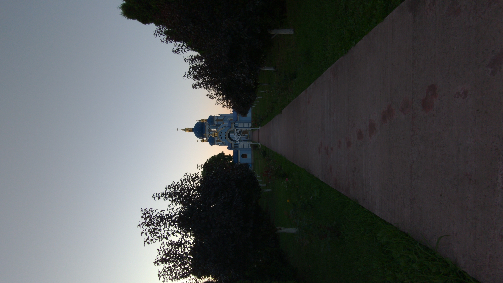 A long concrete path leads towards a light blue and gold domed church, possibly St. Michael's Church, in Zalishchyky, Ukraine. The path is flanked by dark green grass and rows of trees with white-painted trunks. The sky is a pale, clear gray, indicating either early morning or late evening. Footprints are visible on the path, suggesting recent human presence.