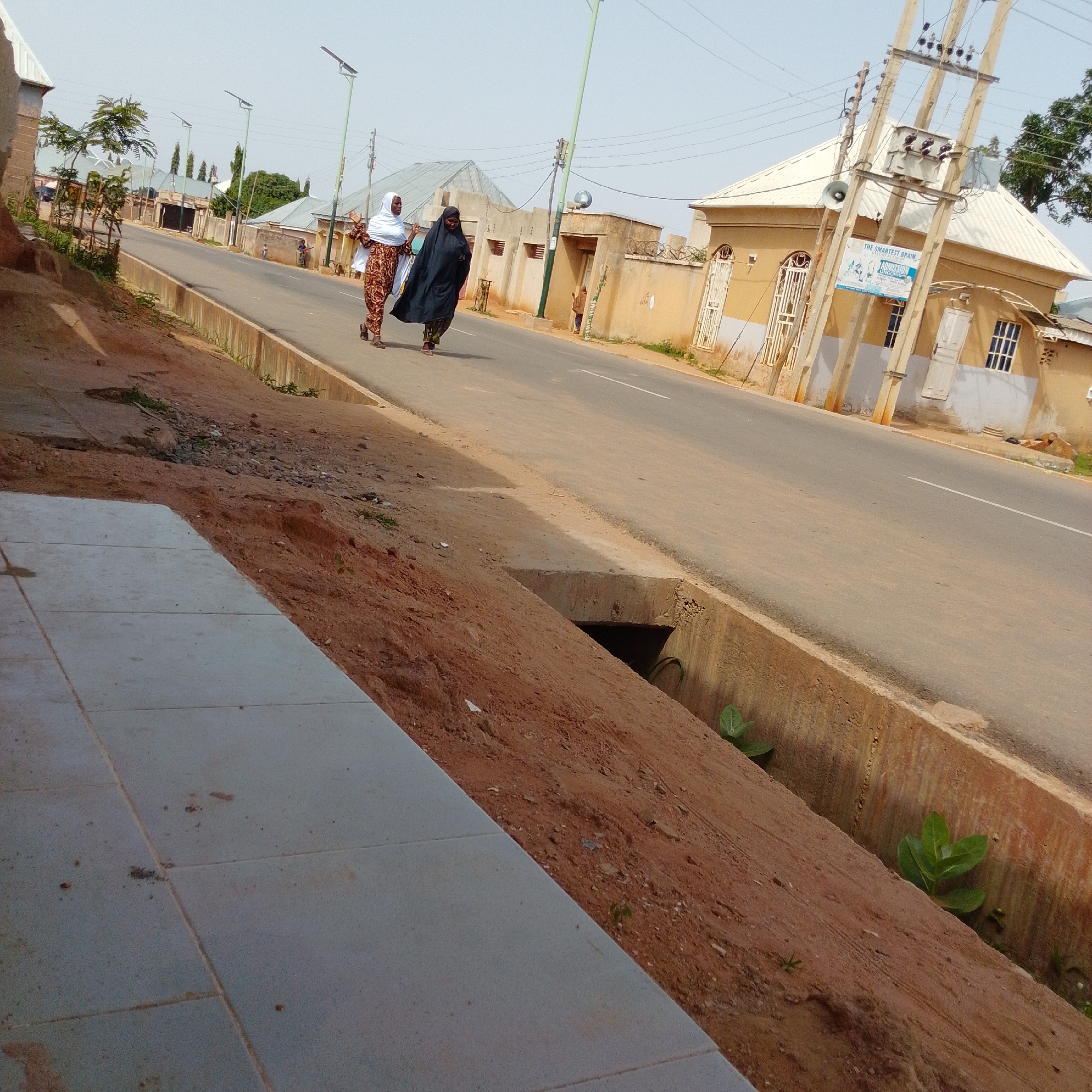 The image depicts a street scene in Gombe, Nigeria. Two women are walking on the right side of the paved road, heading away from the camera. The woman in front is wearing a black abaya and headscarf, while the woman behind her is in a patterned dress and white headscarf, carrying an object in her left hand. A utility pole with electrical wires and a transformer stands prominently on the right side of the road. Adjacent to the road is a dirt embankment with sparse vegetation and a concrete drainage ditch. Several buildings with light-colored walls and corrugated metal roofs are visible along the street. The sky is clear and the lighting suggests it is daytime.