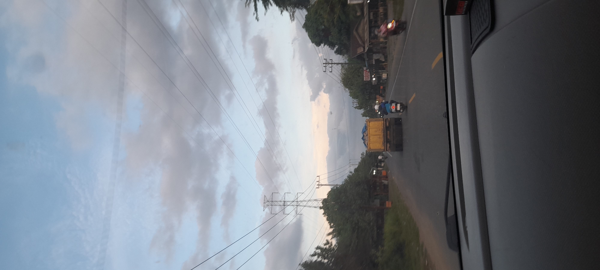 The image shows a view from inside a vehicle, likely a bus or truck, on a paved road. The location is near Paku, Indonesia, inferred from visual cues and the image's origin.
The focus is on the road ahead, where a yellow truck is traveling in the same direction, followed by a motorcycle. A red motorcycle is seen in the distance, moving in the same direction. The road is marked with a yellow line.
To the left of the road, power lines and utility poles are visible against a cloudy sky. Vegetation is present on the roadside. The time of day appears to be dusk or early evening.
The interior of the vehicle is visible on the right side of the image, showing part of the vehicle's wall and a possible window frame.