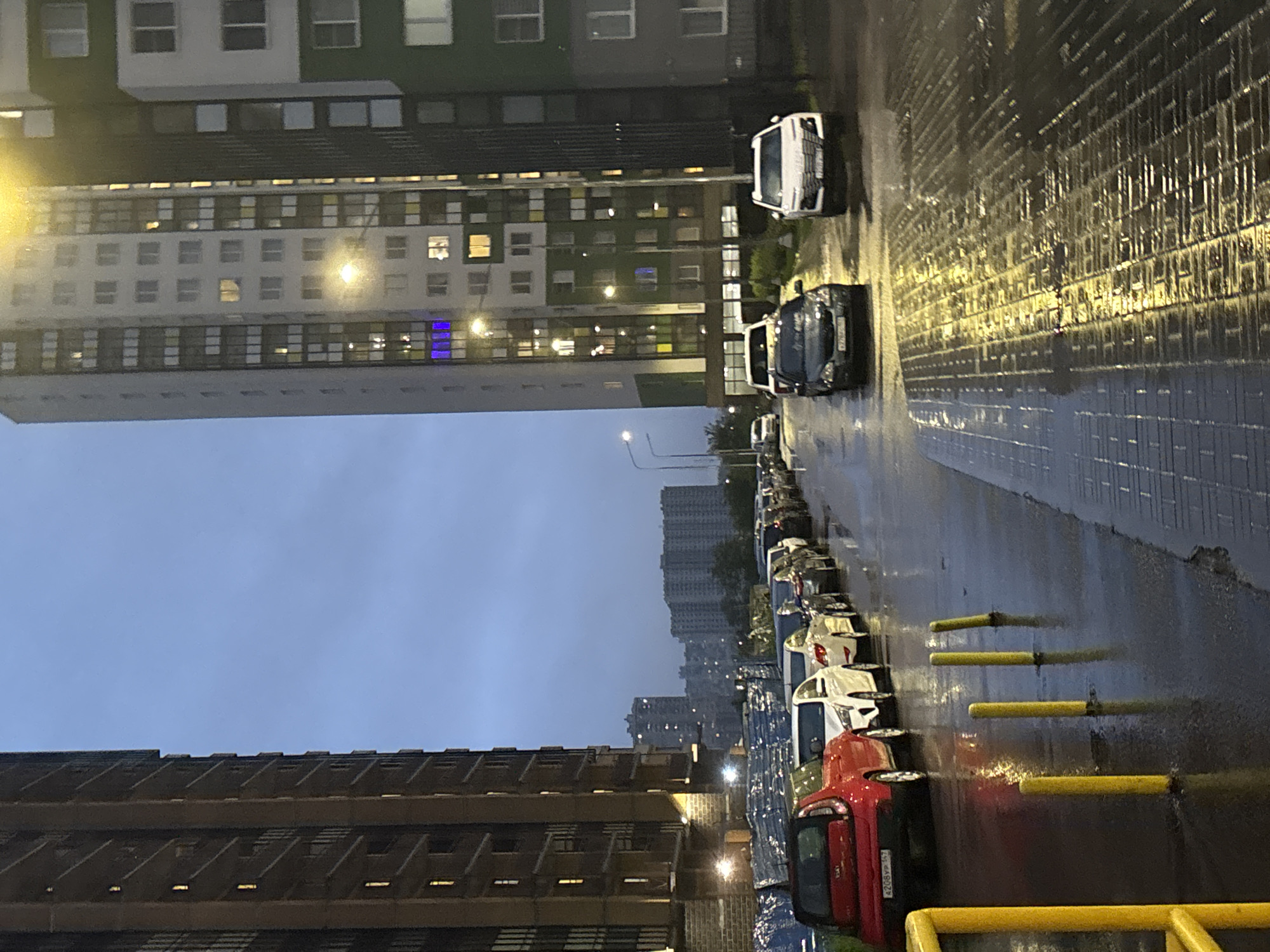 The image depicts an exterior scene in an urban environment, likely during dusk or early evening. Tall, modern apartment buildings dominate the background. The building on the left is a brown, multi-story structure with many windows and balconies. To its right, a taller, predominantly white building with green accents features numerous lit windows. A long line of parked cars stretches along the wet street and sidewalk in the middle ground, reflecting the ambient light. The ground is wet, indicating recent rain or damp conditions, with puddles visible on the pavement. Several yellow bollards are positioned along the sidewalk. In the foreground, a yellow barrier is partially visible on the left. The sky is overcast and a pale blue.