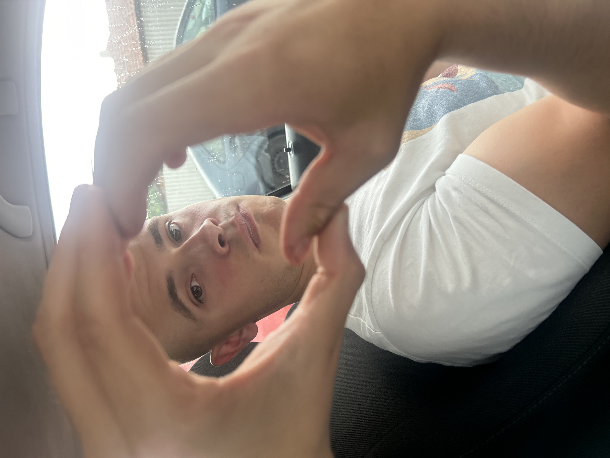 A person with short brown hair is in a car, making a heart shape with their hands in front of their face. The person is wearing a white t-shirt and is seated in a black car seat. The background through the car window shows light-colored surroundings. There is no visible indication of this image being in Elhovo, Bulgaria.