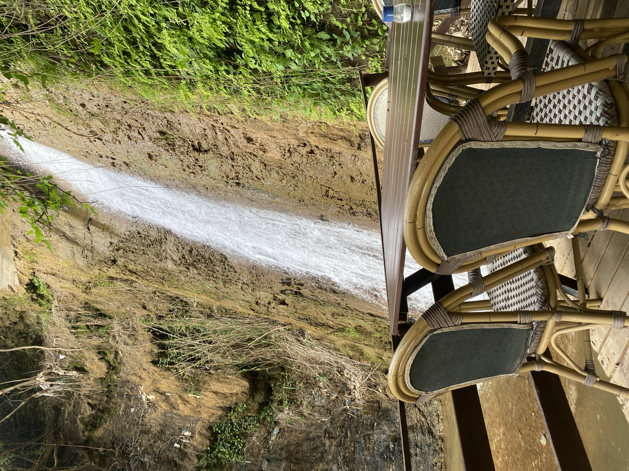 The image depicts a waterfall cascading down a cliff face in Alanya, Turkey. Adjacent to the waterfall is an outdoor seating area with a table and two chairs. The chairs are made of woven material with dark green cushions. A bottle of water sits on the table. The cliff face is covered with greenery and bare patches of soil. The waterfall appears to be the main subject, with the outdoor seating area positioned to allow a view of it. The environment is natural, with the sound of the waterfall likely adding to the ambiance.