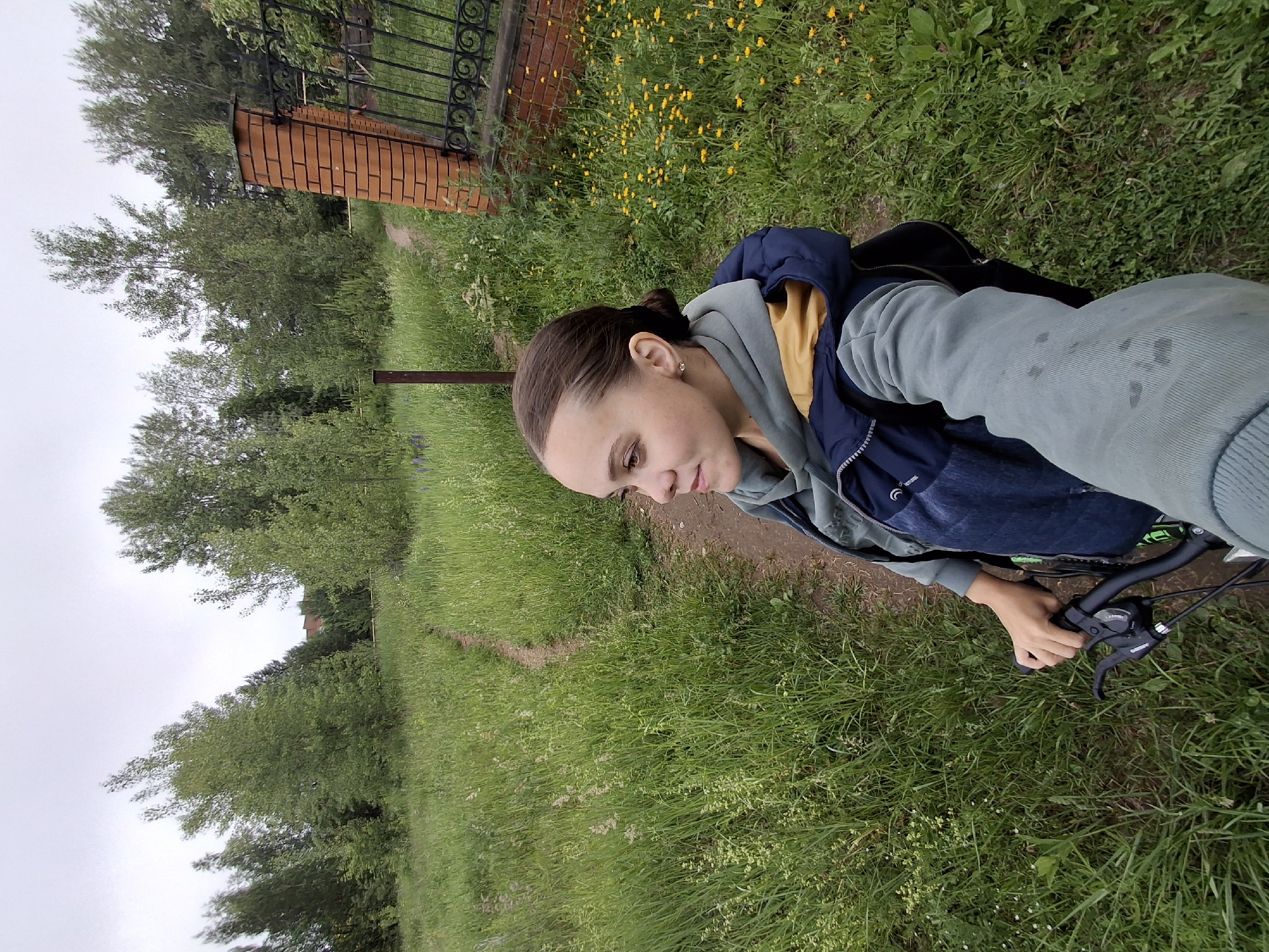 The image depicts a young woman with her hair pulled back in a bun, taking a selfie while holding the handlebars of a bicycle. She is wearing a light blue hoodie layered over a darker blue vest with a yellow collar. The setting appears to be a rural or suburban area with a dirt path winding through tall green grass. In the background, there are trees and a brick fence with an ornate black metal gate. A few yellow wildflowers are visible in the grass near the fence. The sky is overcast, suggesting a cloudy day. The image does not contain any specific landmarks or features that would allow for precise localization to the city of Chaikovsky, Russia.