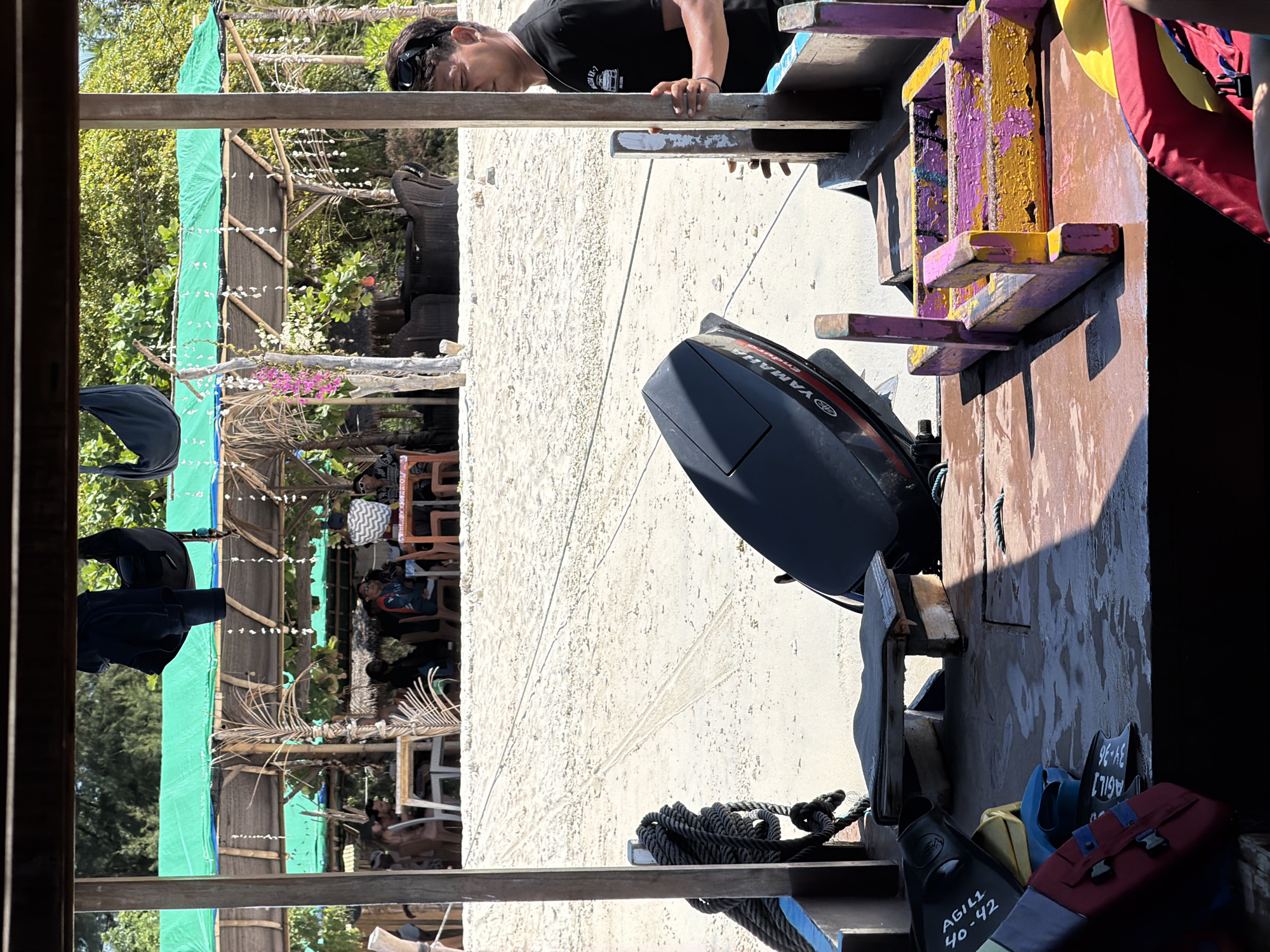 The image depicts a view from a wooden boat moored on a sandy beach. The boat's interior is visible, with fins, life vests, and a Yamaha outboard motor in the foreground. Beyond the boat, a beachside restaurant or seating area with thatched roofs and green coverings is present, with several people seated at tables. Palm trees and lush vegetation form the background. A man wearing sunglasses and a black t-shirt stands on the right side of the boat, looking towards the beach. The overall scene suggests a tropical beach destination.