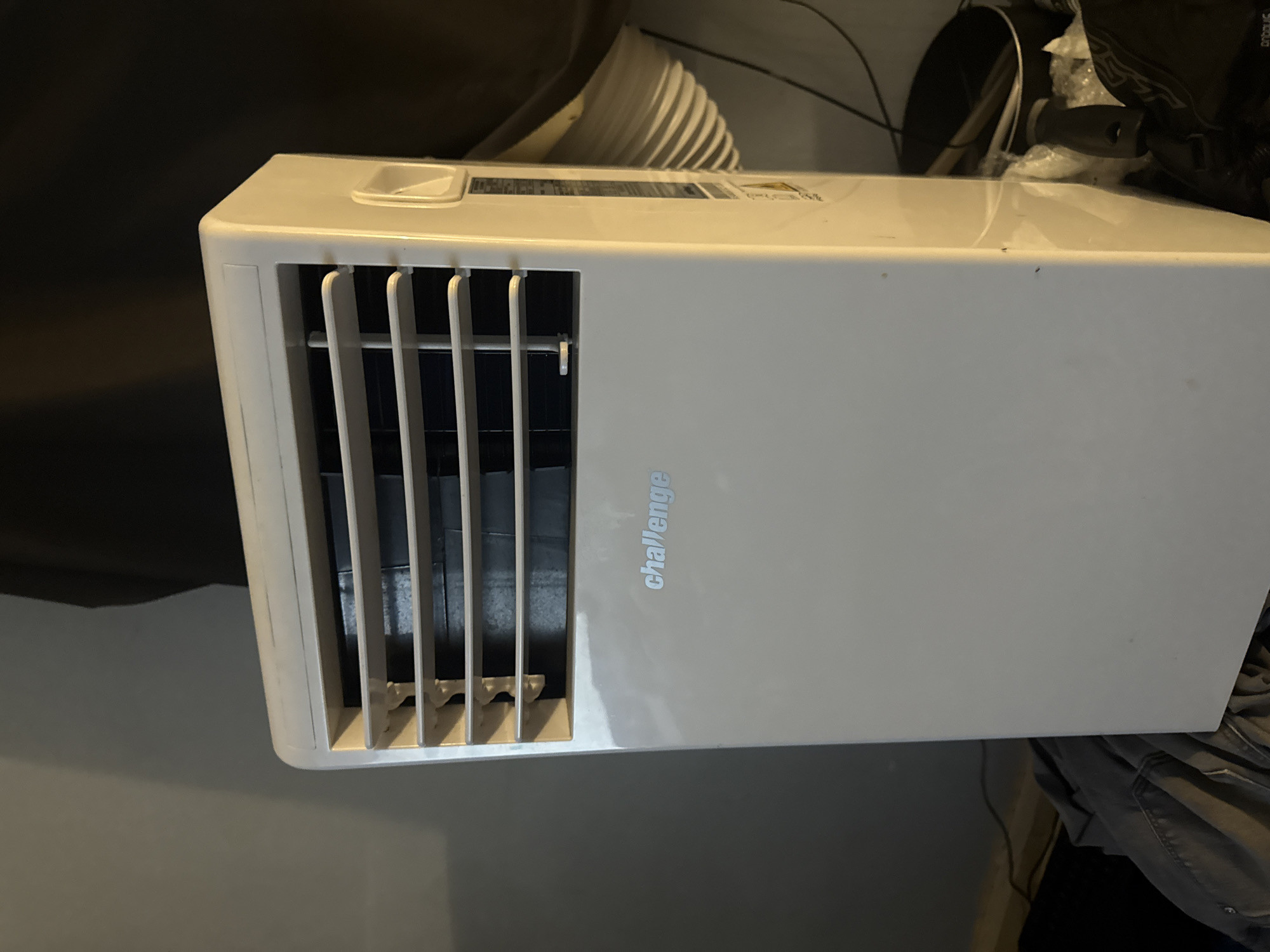 The image shows a white portable air conditioning unit with the brand name "Challenge" visible in silver lettering. The unit has horizontal louvers at the top, suggesting it is designed to expel air. A white flexible duct is attached to the side and curves out of view, indicating it is likely connected to a window or vent. The air conditioner is positioned in an indoor setting, against a grey wall on the left and a dark background on the right. In the foreground, on the left side of the image, there are some dark grey garments, possibly clothing, and a black electrical cord. On the right side, towards the bottom, there are various household items, including a black pot and some plastic bags. The lighting in the scene is dim, with shadows cast on the wall and the unit itself. The image does not contain any people or evidence to suggest a specific location within the city of Cheltenham, United Kingdom, beyond it being an indoor domestic environment.