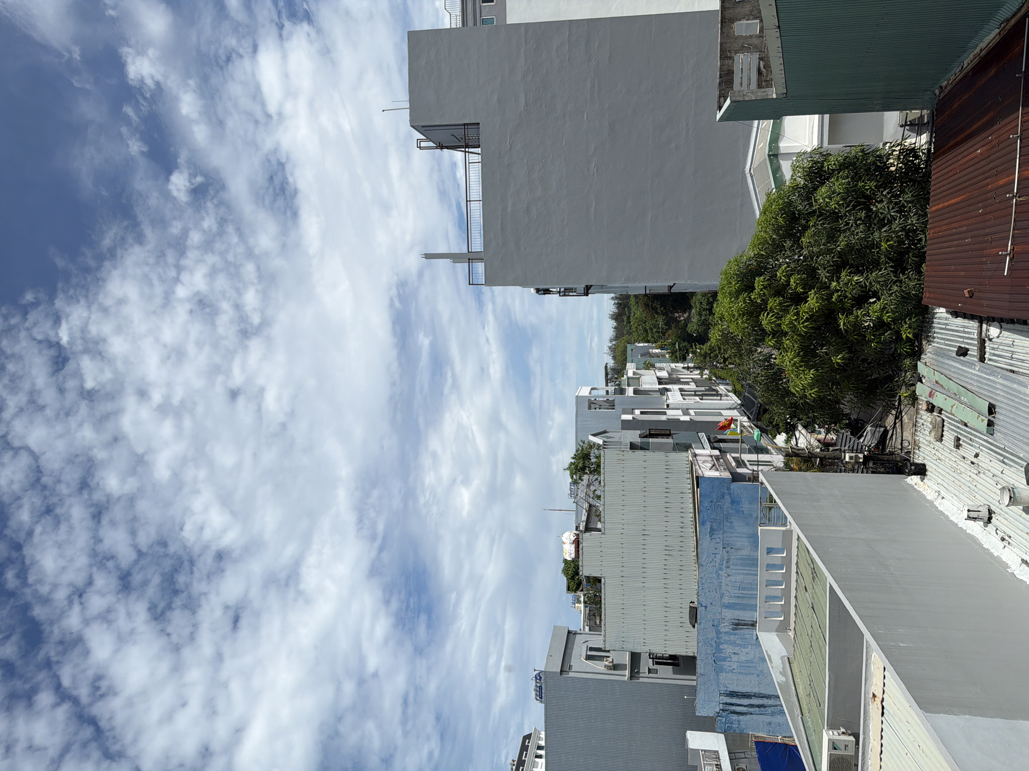The image is an outdoor, elevated view of a neighborhood in Đà Nẵng, Vietnam. Predominantly clear sky, scattered with cumulus clouds, occupies the upper left section. The neighborhood is densely packed with buildings of varying heights and architectural styles. The buildings have a mix of modern and older designs. The color palette is muted, with shades of gray, white, blue, and green dominating the scene. A mature tree is visible, adding a natural element to the urban landscape. Corrugated metal roofs are present, indicating the tropical climate. There are no visible people in the image.