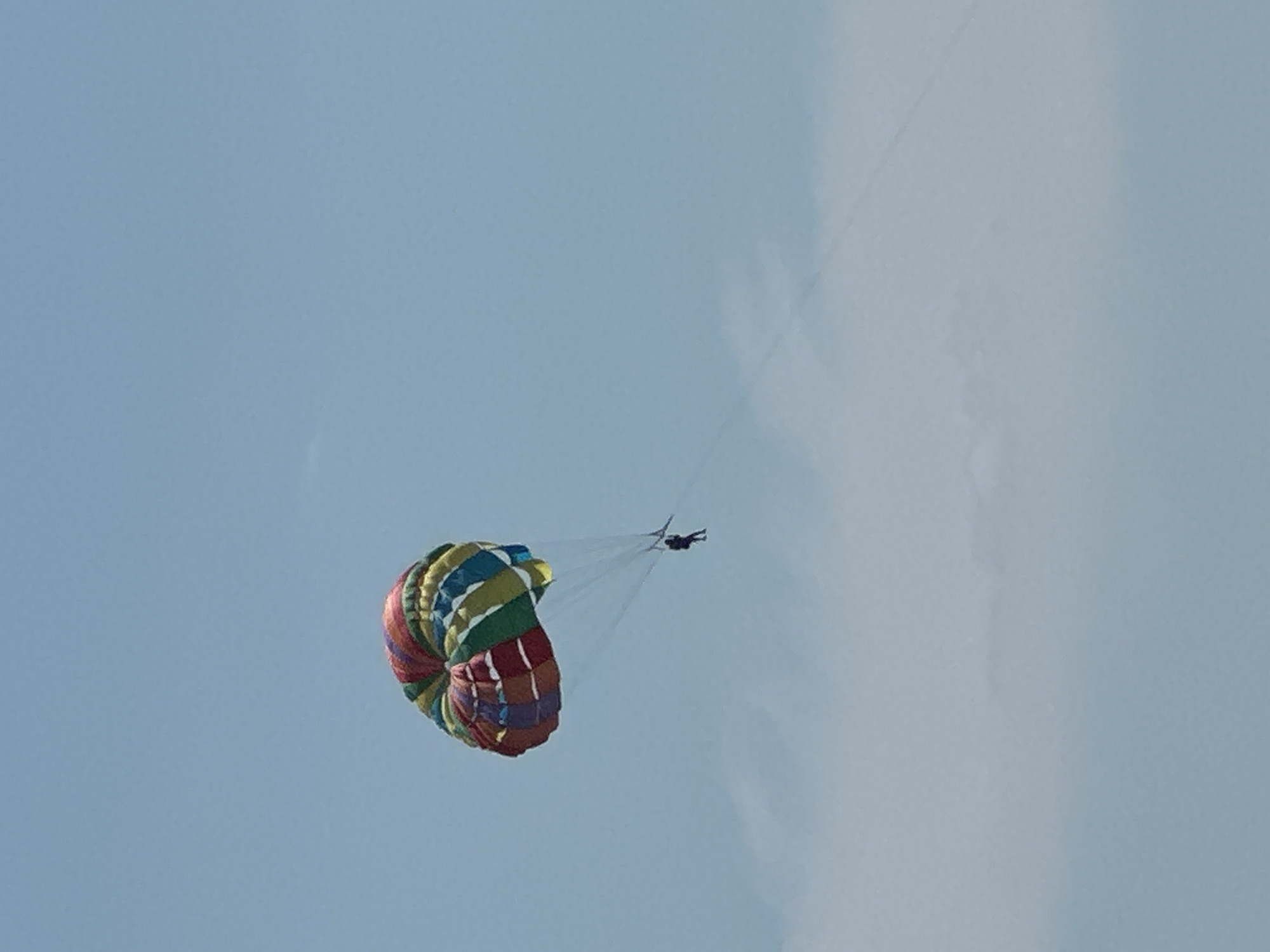 A single person is parasailing in a clear blue sky. The parasail is colorful, with alternating stripes of red, yellow, green, and blue. The person is suspended below the parasail, attached by lines. In the background, there are some wispy white clouds. The location is the city of Boreti, Montenegro.