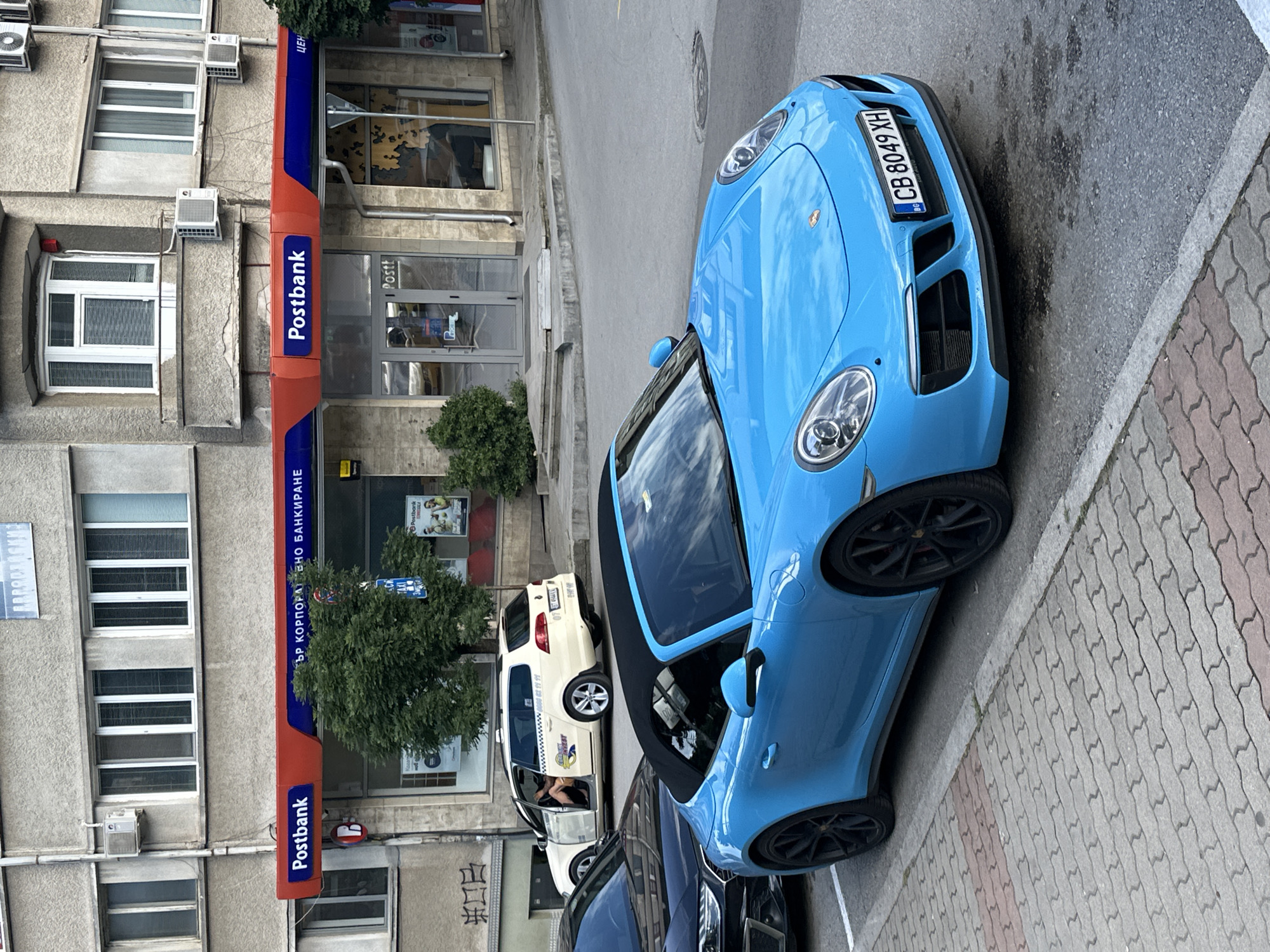 A bright blue convertible Porsche is parked on a city street in Veliko Tarnovo, Bulgaria. The car is in the foreground, facing right, with its black soft-top down. The license plate reads "CB 8049 XH". Behind the Porsche, a white taxi van with the door open is parked, and a person's legs are visible inside. Further in the background, a Postbank branch is visible with a red and blue awning and large glass windows. The building appears to be a multi-story structure with numerous windows. The street is paved with cobblestones in the foreground and asphalt further back. There are trees and foliage visible in front of the Postbank. The overall environment suggests an urban setting.