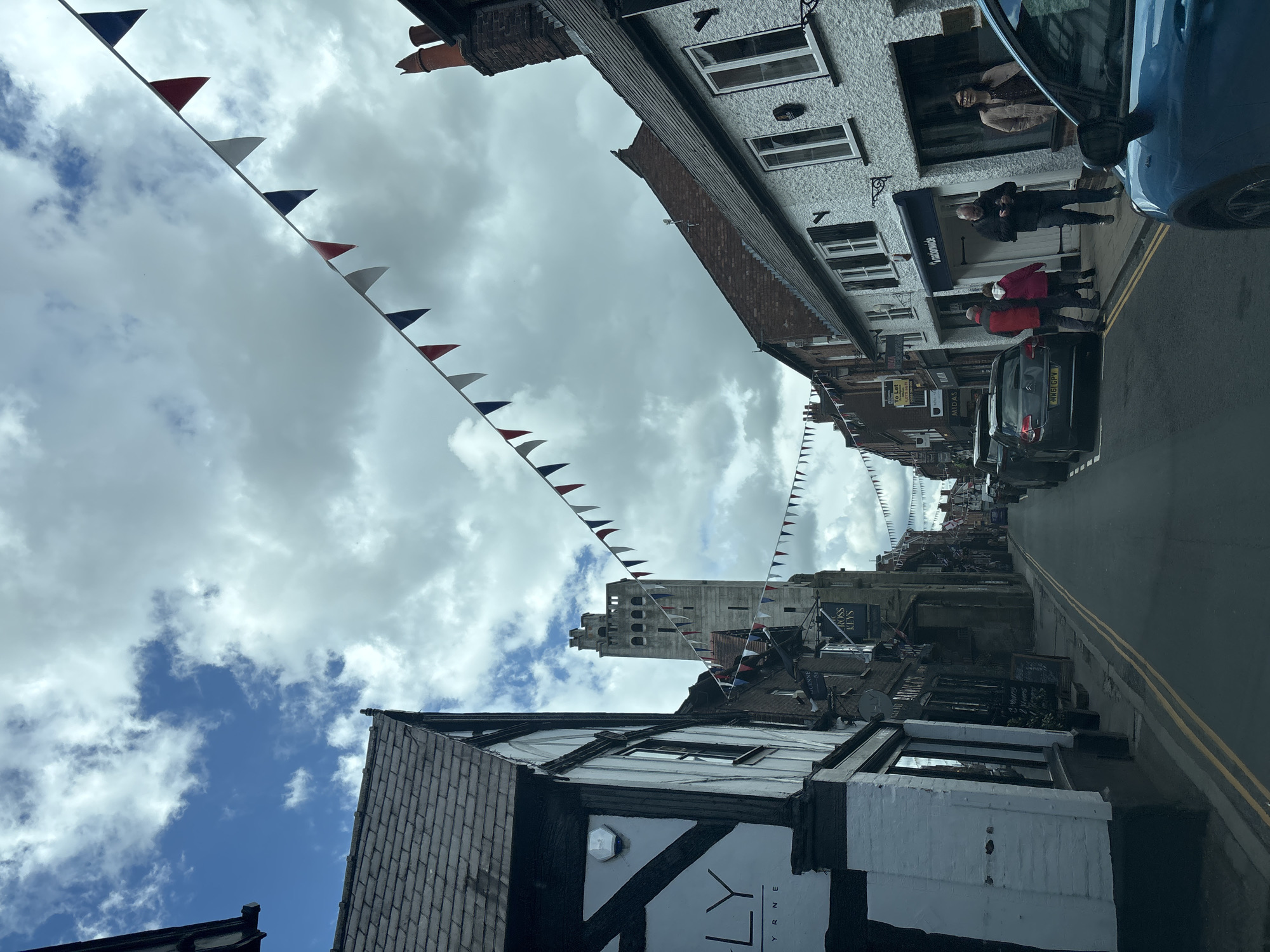 The image shows a street view in Knutsford, United Kingdom. The street is lined with buildings, some of which are Tudor-style with black and white facades. Festive bunting in red, white, and blue hangs across the street. There are cars parked on the side of the road. Several people are walking along the pavement. A tall, stone tower can be seen in the distance at the end of the street. The sky is cloudy, with patches of blue. Double yellow lines are visible in the center of the road.