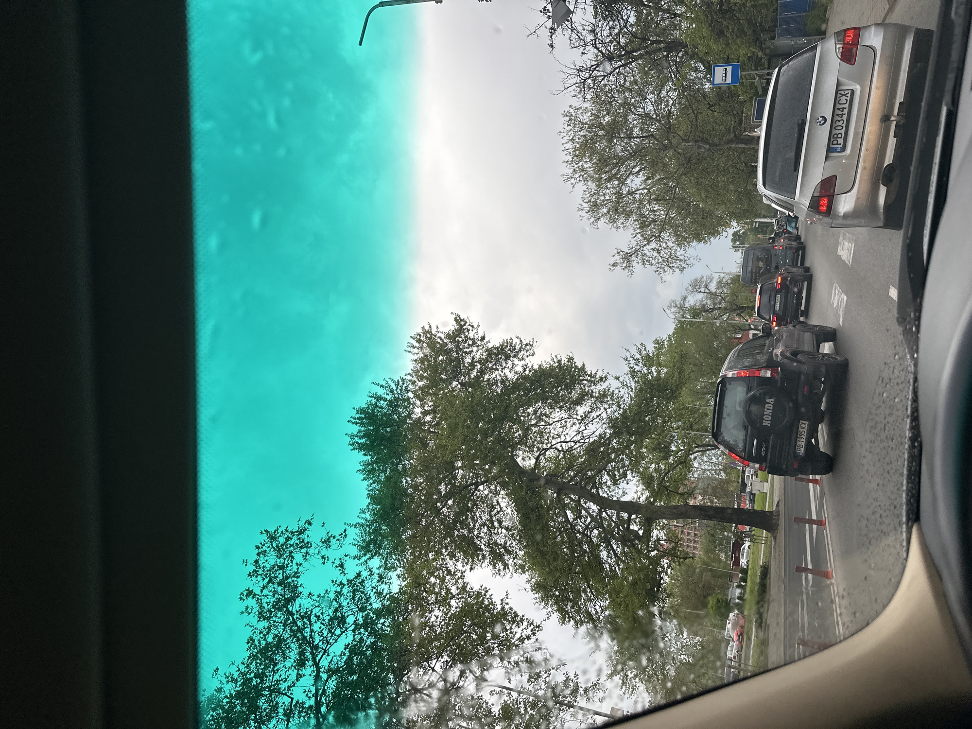 The image, captured from inside a vehicle, presents an urban street scene in Plovdiv, Bulgaria, viewed through the windshield. The upper left portion of the windshield exhibits a distinct teal-colored tint, possibly from a sunshade or built-in tinting.
The scene depicts a street with several cars in traffic. Most prominently, the rear of a silver BMW station wagon is visible, displaying the Bulgarian license plate "PB 0344 CX," where "PB" indicates the Plovdiv region. Further ahead in the queue, a black Honda CR-V is seen from behind, with "HONDA" and "CRV" badging, and another Plovdiv license plate "PB 1995 KX." On the right side of the road, near the Honda, several orange traffic cones are placed.
The street is lined with numerous deciduous trees, featuring lush green foliage, suggesting a spring or summer setting. The sky above is overcast or partly cloudy. In the background, a blue bus stop sign, typical of European cities, is visible on the left. The windshield shows some streaks or moisture, hinting at recent rain or damp conditions. The overall impression is a busy daytime city street with ongoing traffic.