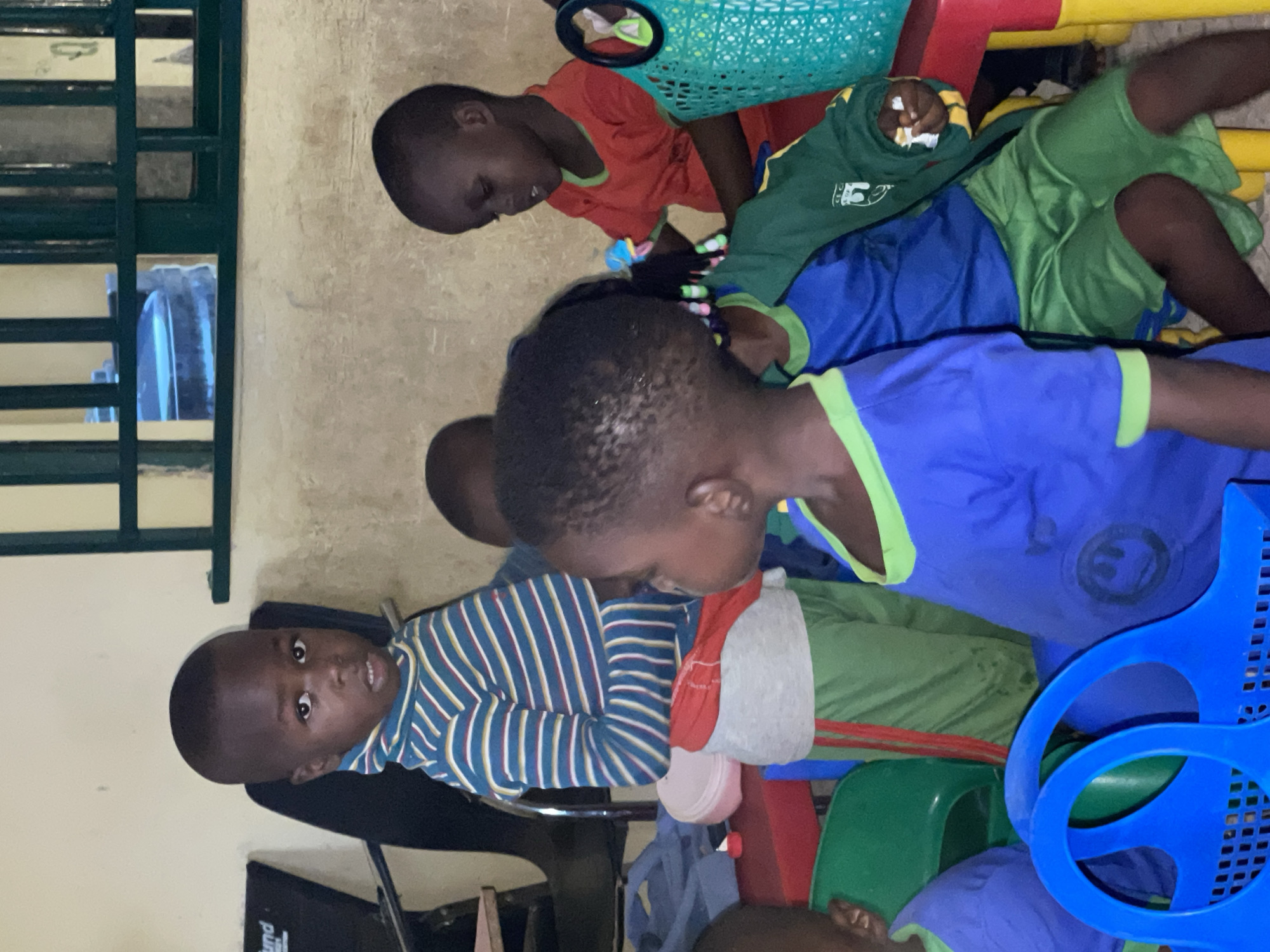An indoor photograph, likely taken in Akwanga, Nigeria, depicts a group of six or more young children, predominantly toddlers or preschoolers, gathered in what appears to be a daycare or classroom setting. The overall lighting is dim, suggesting either evening or a poorly lit interior.
In the foreground on the left, a young boy wearing a blue and white striped long-sleeved shirt sits on a dark chair, looking directly at the camera with a neutral, slightly curious expression. To his right, another child in a blue short-sleeved shirt with lime green trim is visible, looking downwards. Several other children are positioned behind and around them. One child wears a similar blue and green uniform, while another in a red short-sleeved shirt is seen near a green plastic basket. A child wearing green shorts and a green long-sleeved top is nestled within a colorful plastic play structure featuring red, yellow, and blue components.
The background consists of light-colored, somewhat textured walls. On the left side of the image, a dark green metal-barred structure, possibly a window with security bars or a room divider, is visible. Through these bars, a blue bottle can be discerned. Additional objects include dark-colored chairs, a grey fabric item (possibly a pillow or blanket), and a large blue plastic object that appears to be part of the play structure or a toy. In the bottom left corner, a portion of a black object, possibly a speaker, shows the partial word "Sound" printed on its surface. The children appear to be in a state of quiet observation or resting.