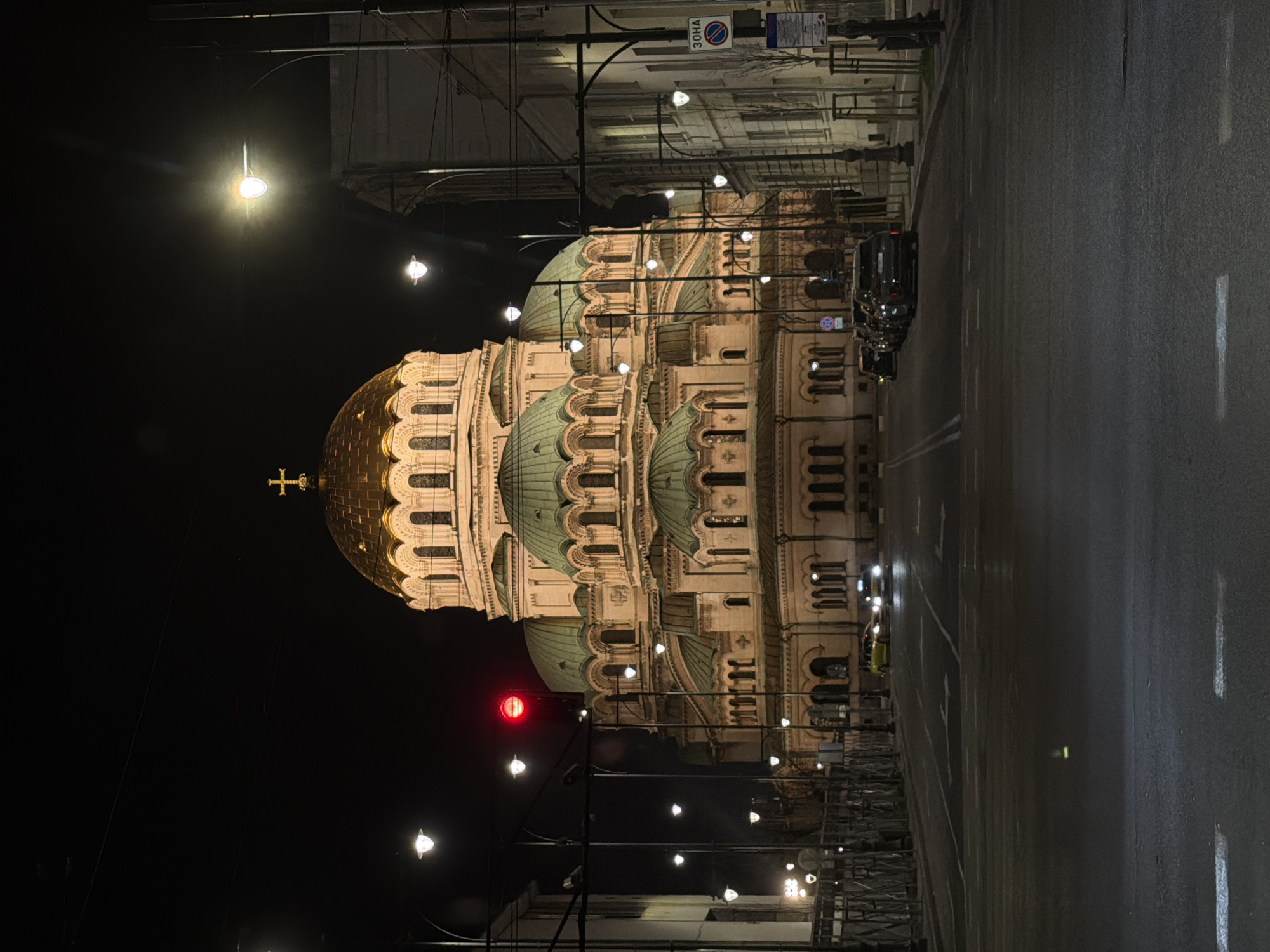 The image displays a nighttime street scene in Sofia, Bulgaria, focused on the Alexander Nevsky Cathedral. The cathedral, centrally framed, features a prominent golden main dome topped with a cross, complemented by multiple smaller green domes and a light-colored stone facade with arched windows, all externally illuminated.
In the foreground, an asphalt road with white lane markings extends towards the cathedral. Two vehicles with illuminated headlights are visible in the middle distance, proceeding towards the cathedral; one appears yellow, the other is dark. A line of dark colored parked vehicles is present on the right side of the street.
The street is lined with numerous illuminated streetlights. A single red traffic light is visible on the left. Utility poles and overhead wires crisscross the scene. Buildings with visible windows and architectural details line both sides of the street. A blue circular 'no stopping' sign with the text 'ЗОНА' (ZONE) is visible on the right. Dark, criss-crossed metal barriers are situated along the left sidewalk, and bare trees line the right. The background consists of a dark, clear night sky. No individuals are visible within the scene.
