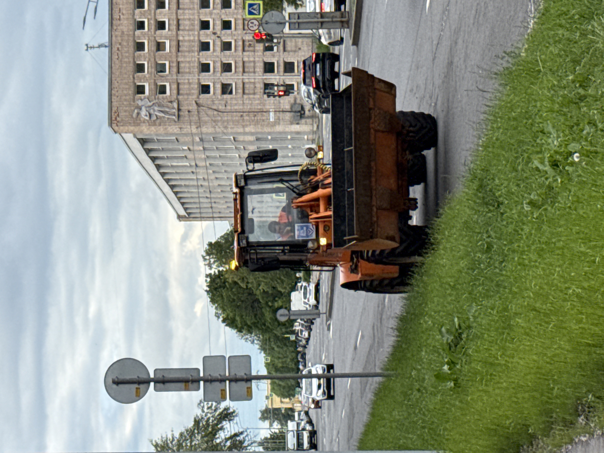 The image shows an orange front-end loader driving on a street in Saint Petersburg, Russia, on a cloudy day. The loader is positioned in the foreground, on the right side of the road, facing away from the viewer. A person wearing an orange vest is visible inside the cab of the loader. Behind the loader, there is a multi-lane road with several cars and trucks. To the left of the road, there is a grassy verge and a pole with traffic signs. In the background, a large, light-colored building with many windows and a bas-relief sculpture on its facade is visible. Traffic lights are also present on the right side of the road. The sky is overcast.