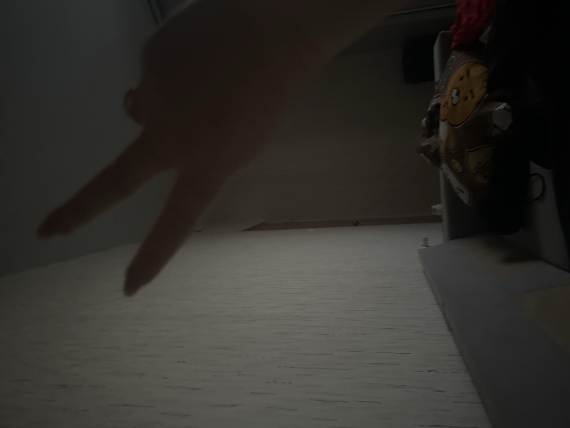 The image captures a dimly lit indoor scene, likely at night or in a poorly lit room. A human hand is prominently featured in the upper left, extending two fingers upwards in a peace or victory sign. The hand is partially illuminated, creating soft shadows.
Below the hand, a light-colored, textured surface stretches across the foreground, resembling wood grain or a patterned floor. To the right, a dark, indistinct object, possibly a piece of furniture or a bag, occupies a significant portion of the frame. This object has a discernible pattern featuring a yellow and brown cartoon-like face. A piece of red fabric is draped over the top of this dark object. Another dark, rectangular shape is visible in the upper right background. There is no visible text in the image. The low light conditions contribute to a subdued atmosphere.