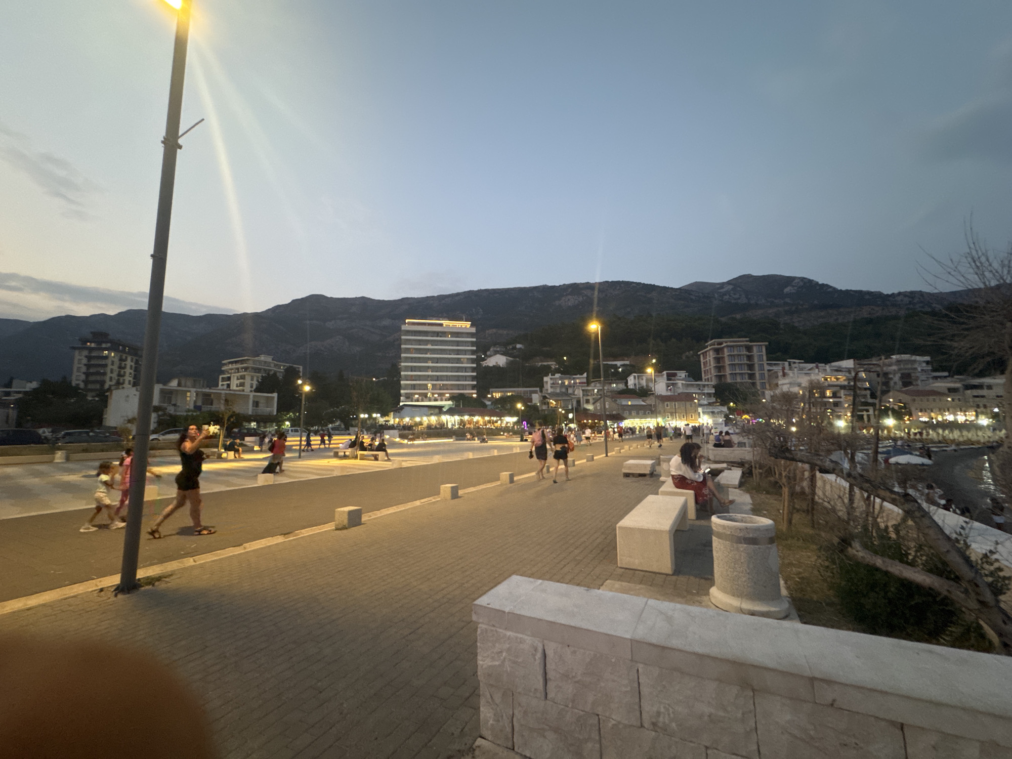 The image depicts a sunset scene along the waterfront promenade in Sutomore, Montenegro. The sky is a clear, soft blue, transitioning into darker hues towards the horizon, with mountains forming a silhouette in the background. The promenade is paved with textured grey stones and lined with a curb and white square bollards. Several street lamps are visible, casting a warm glow on the scene.
On the promenade, several individuals are present. In the foreground, two young girls, one in a pink top and white shorts, the other in a white top and pink skirt, are walking away from the camera. To their right, a woman in a dark dress and sandals is walking, holding a phone. Further down the promenade, other people are seen walking and sitting on benches. Some are gathered in groups, while others appear to be relaxing or enjoying the evening. Buildings line the background, illuminated by artificial lights, suggesting a commercial or residential area. The sea is visible on the right side of the image, with a beach and some structures along the shore. The overall atmosphere is one of a calm evening stroll along a coastal town.