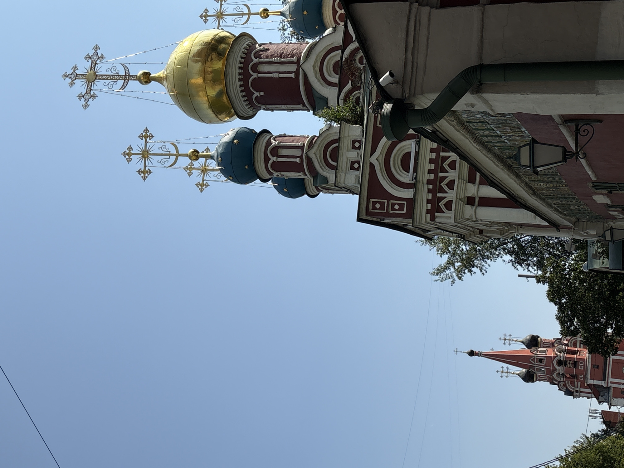 This is a low-angle, outdoor photograph of multiple domed churches against a clear blue sky. The dominant church features several domes: one large, golden dome, and two smaller, blue domes. The architecture is red and white with intricate detailing. Another church, predominantly red with black domes, is visible in the background on the left, appearing smaller due to distance. The image captures religious structures, suggesting a religious or historical site. The presence of trees and a lamppost implies an urban setting. The photograph does not contain any people or overt actions. The scene is static and focused on the architecture.