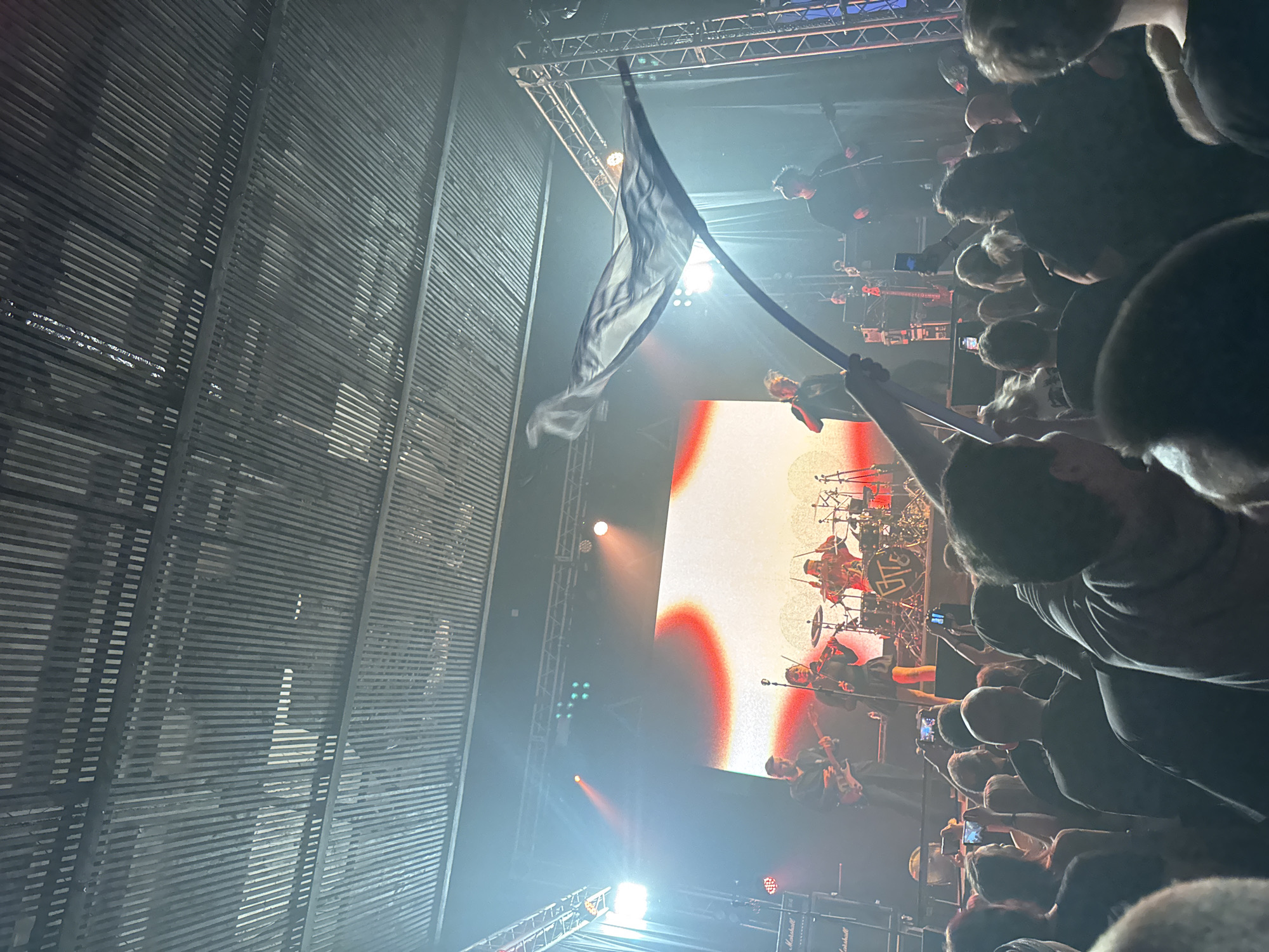 A concert is being held in Izhevsk, Russia. A person in the audience is holding a flag. The band is performing on stage in front of a large screen. The audience is recording the concert on their phones.