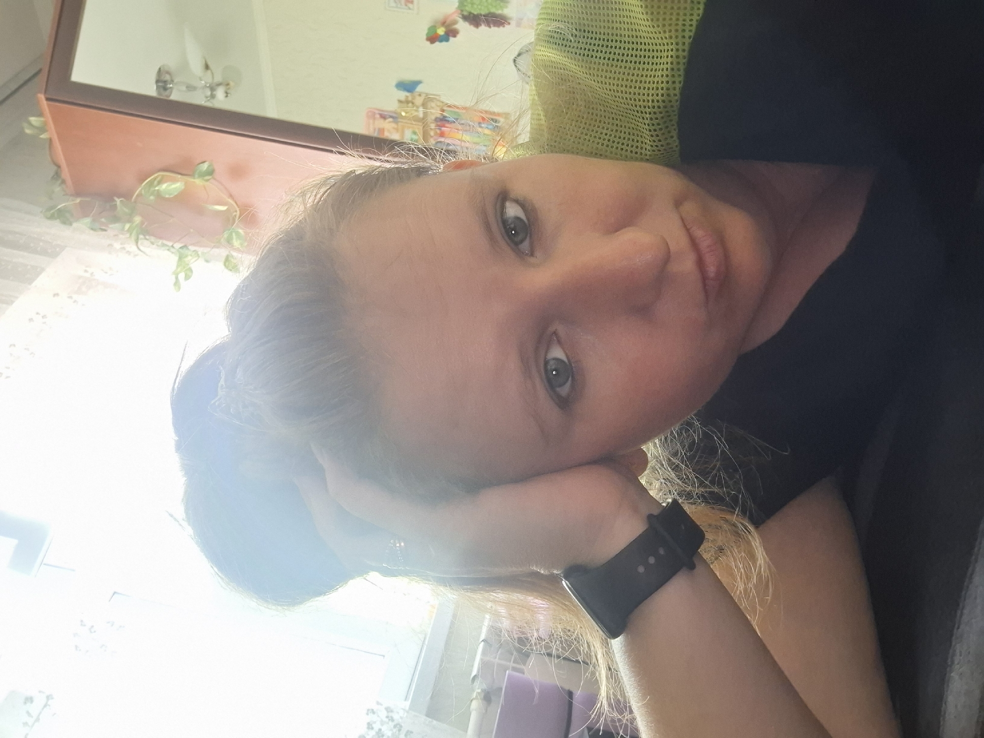 The image shows a close-up selfie of a woman with her right hand resting on her forehead and temple, her hair pulled up in a messy bun. She is wearing a black t-shirt and a black smartwatch on her left wrist. The background is blurred but appears to be an indoor setting, possibly a room with a window covered by lace curtains. A wooden cabinet with a hanging plant is visible to the right, along with a glimpse of a ceiling fan and a mirrored surface reflecting more of the room. The lighting suggests it might be daytime, with sunlight streaming in from the left. There are no visible indicators of the specific location within the city of Shymkent, Kazakhstan.