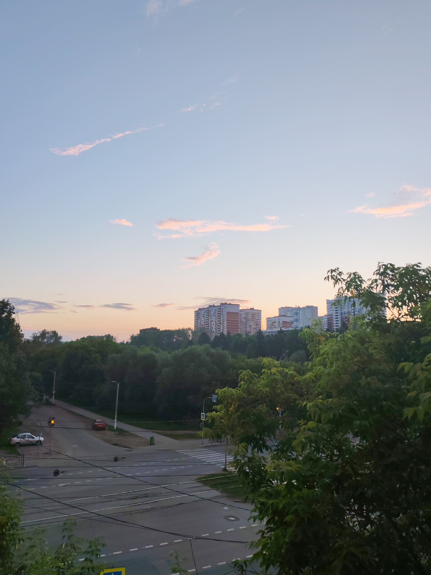 The image depicts a street intersection in Izhevsk, Russia, during a sunset. The sky is a soft gradient of blue and pink, with scattered clouds. In the foreground, lush green trees partially obscure the view. The intersection itself features asphalt roads with visible lane markings and pedestrian crossings. A traffic light at the intersection shows a yellow signal. Two cars are present: a silver car parked on the left side of the road and a red car driving away from the camera. Several utility poles and overhead wires are also visible. In the background, a dense line of trees separates the intersection from a cluster of apartment buildings. The buildings are of varying heights and architectural styles, suggesting a typical urban residential area. The overall scene conveys a tranquil, late-day urban atmosphere.