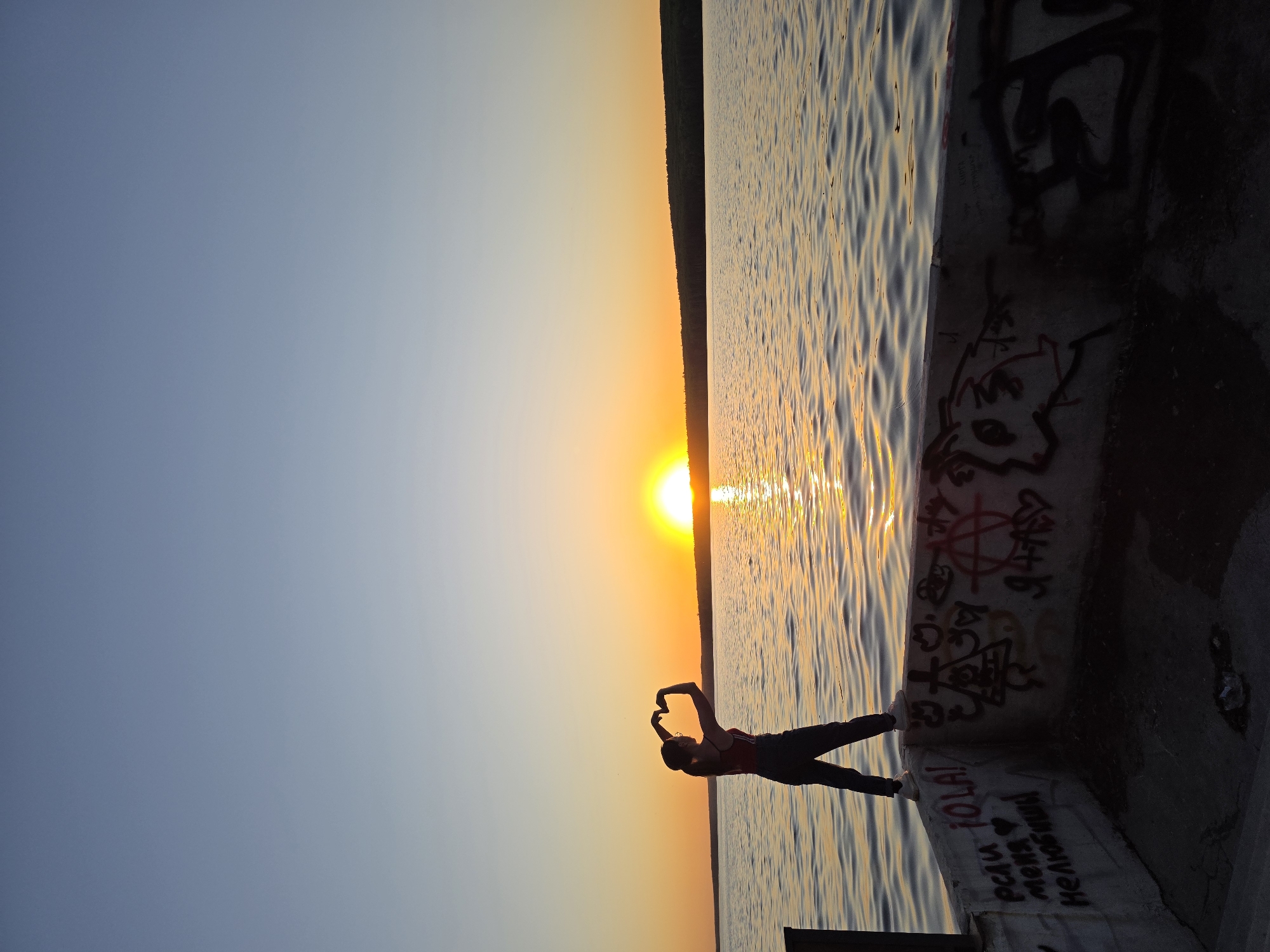 A silhouetted figure stands on a concrete pier at sunset, with graffiti on the pier's edge. The figure makes a heart shape with their hands towards the sun. The water reflects the sun's light, and the background shows a dark treeline. The graffiti includes Cyrillic text and symbols. The location is Chaikovsky, Russia.