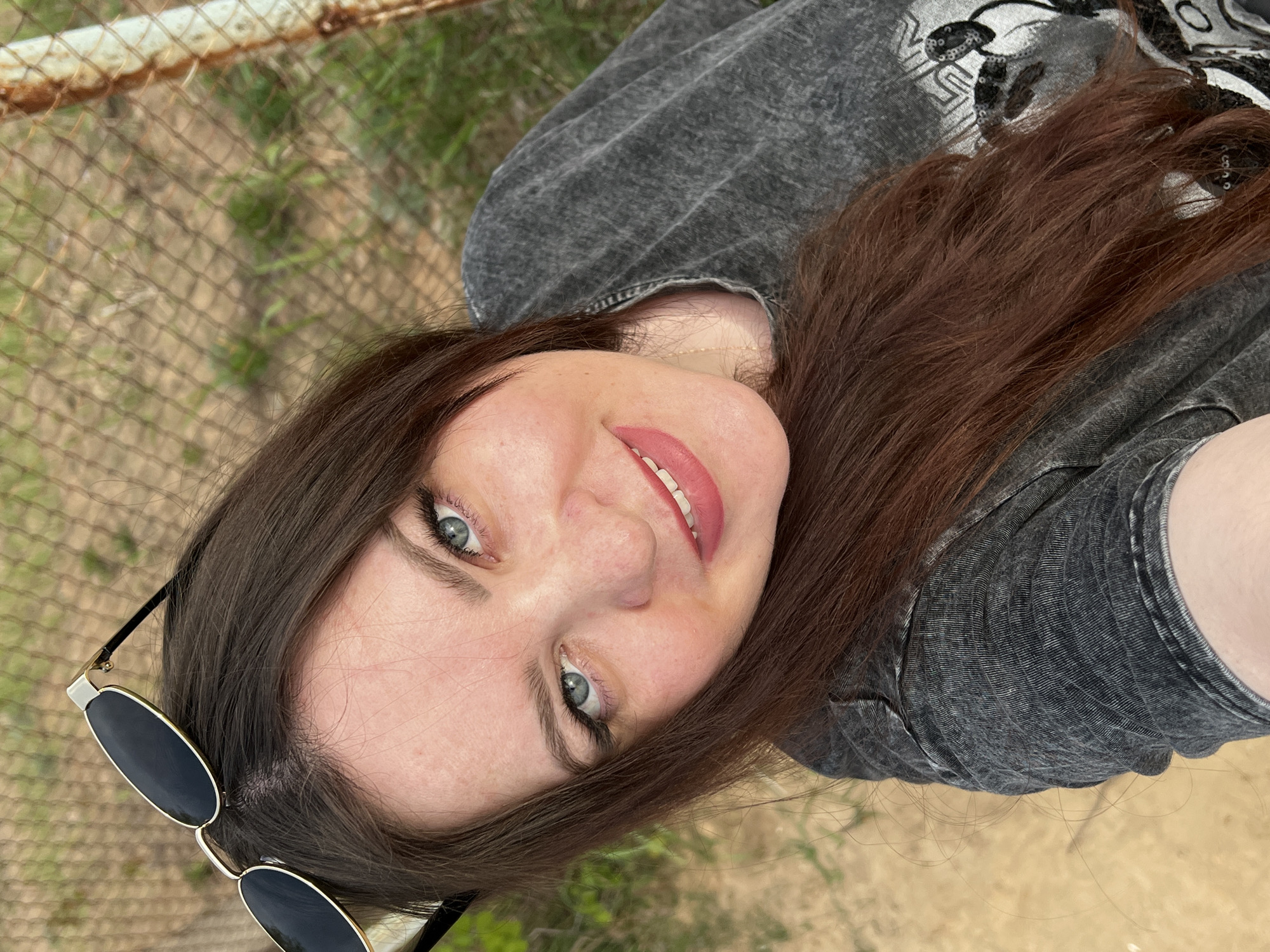 A woman with long brown hair and light eyes is taking a selfie. She is wearing a gray shirt and has sunglasses perched on the side of her head. The background consists of a rusty wire fence and some greenery. The location is unknown, but the prompt suggests it might be in the city of Noviy, Russia.