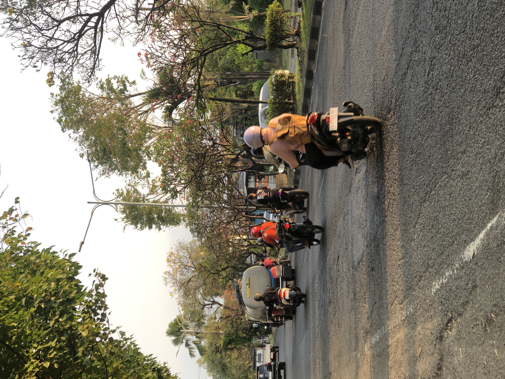 The image depicts a street scene in Gelam, Indonesia, during daylight. Several motorcycles are in motion on a paved road. One motorcycle in the foreground has a rider wearing a pink helmet and a yellow backpack. Further back, a motorcycle rider in an orange helmet is visible. A large tanker truck is positioned in the middle ground, with several other motorcycles and a white pickup truck also present on the road. The environment features trees with lush green foliage and some flowering trees with pink blossoms. A street lamp is visible in the upper left portion of the image. The sky is bright and appears to be a pale yellow or white, suggesting either early morning or late afternoon light. The road surface is asphalt with some visible markings and texture. Buildings and vegetation line the sides of the road.