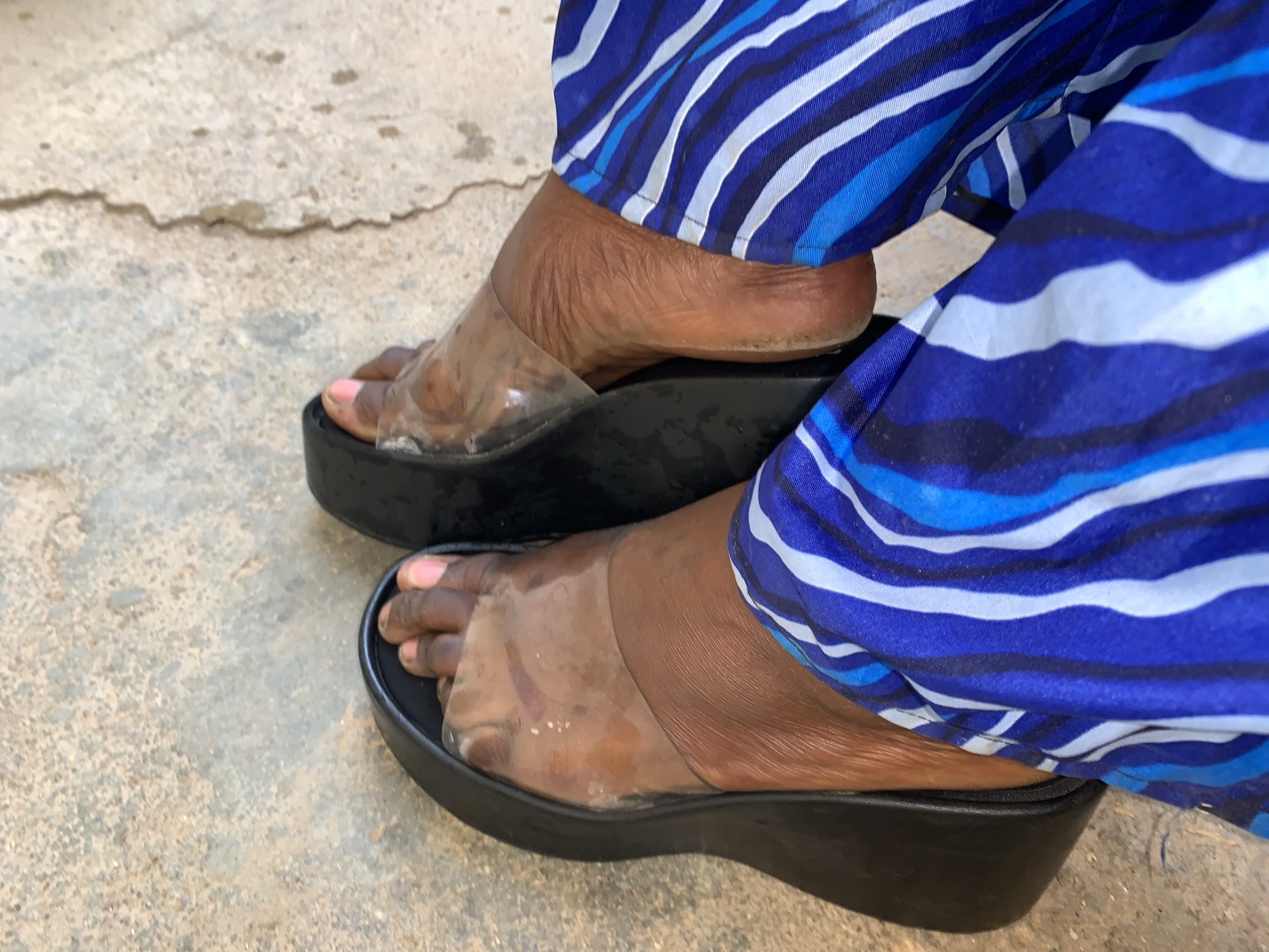 An image shows two feet clad in clear plastic sandals with black soles, against a concrete surface and blue and white striped fabric, in an outdoor setting. The location is implied to be Rafin Par, Nigeria. The feet are in the foreground, positioned close together, with the toes and insteps visible under the clear plastic straps. The sandals are wedge-heeled. The person wearing the sandals is also wearing loose, blue and white striped pants or a skirt, with the fabric draped around the ankle area. The backdrop is a plain, cracked concrete surface.