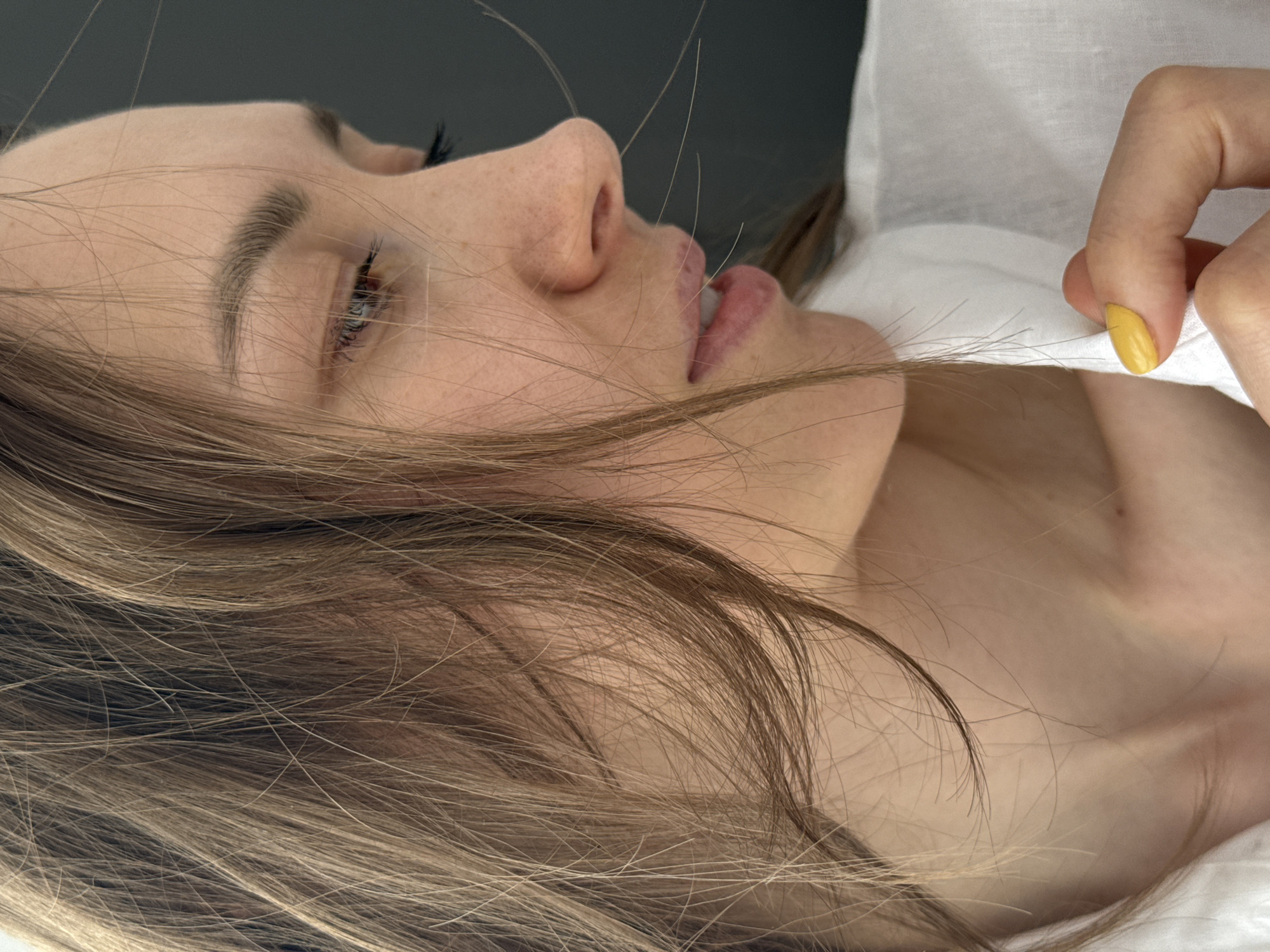 A close-up image shows a fair-skinned woman with light brown hair lying on her side. Her eyes are closed and her lips are slightly parted. Her hair is draped across her face and neck. A hand with yellow nail polish is visible in the upper right corner. The background is a mix of dark grey and white. There is no evidence to suggest this image was taken in Chaikovsky, Russia.