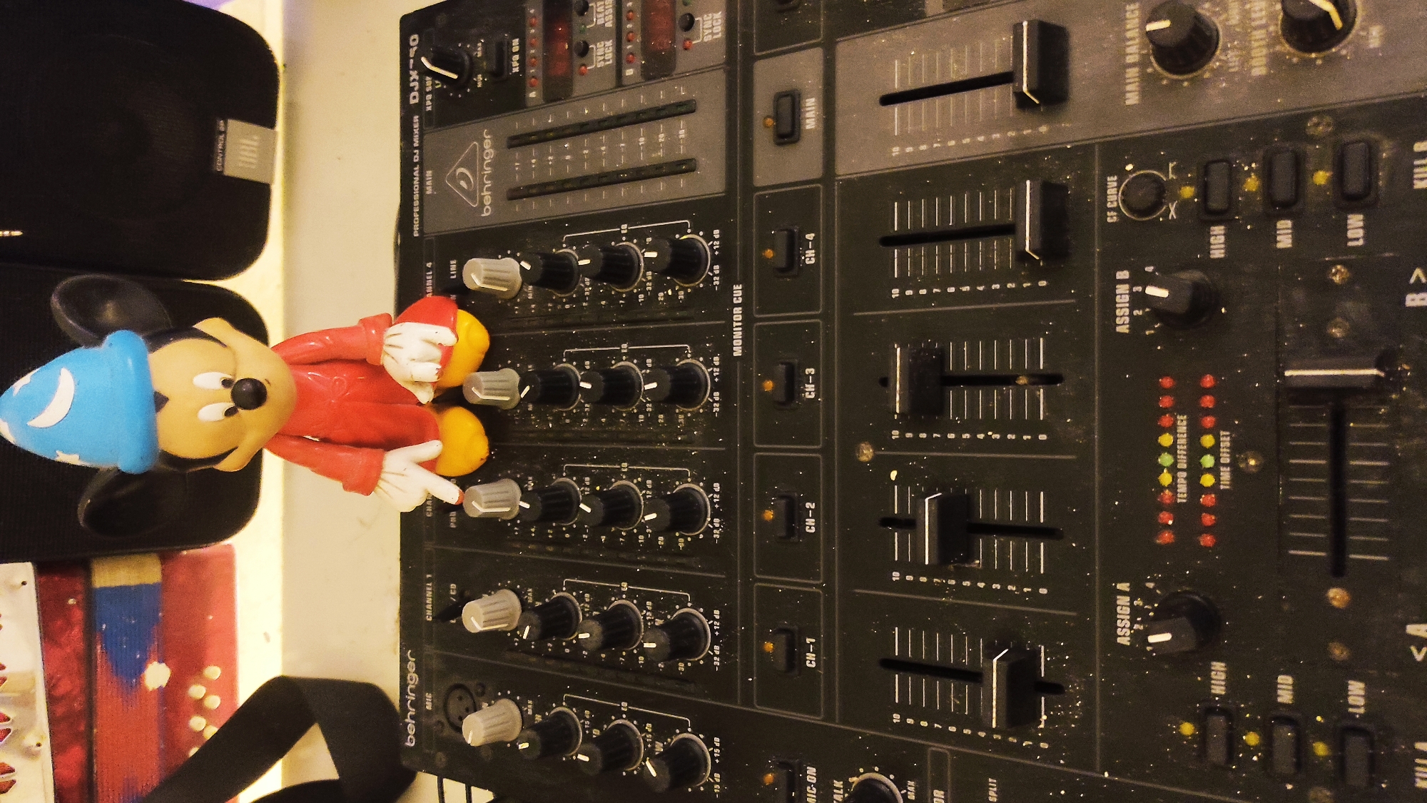 A Mickey Mouse figurine wearing a blue wizard's hat and a red outfit stands on a Behringer DJ mixer. The mixer is black and has several knobs, faders, and buttons. The figurine is positioned in the center of the mixer, behind the knobs for channels 2 and 3. Dust is visible on the mixer's surface. In the background, two black speakers are partially visible. The overall scene appears to be indoors, possibly a room or studio. The location of Remedios de Escalada, Argentina is not depicted in this image.