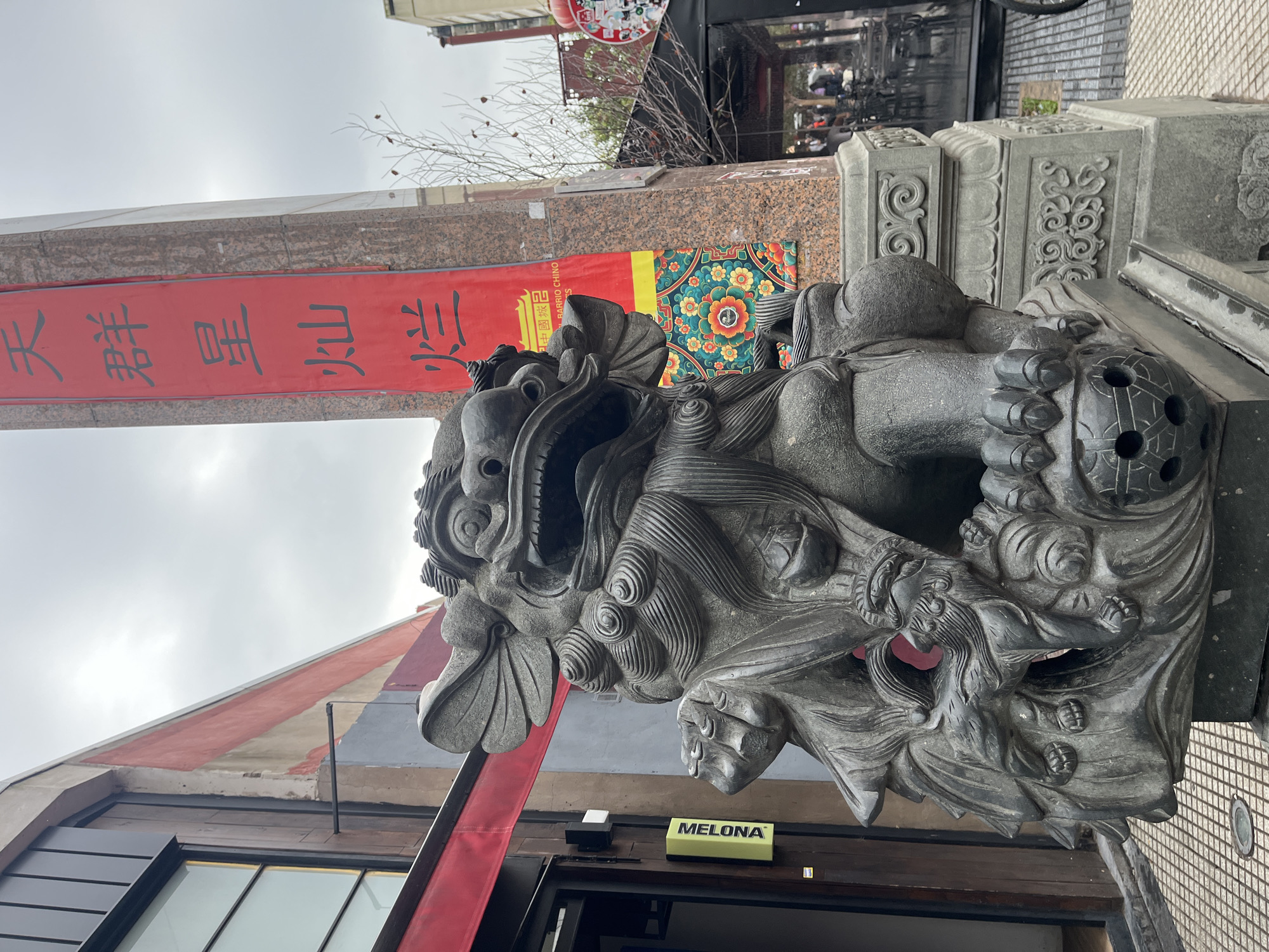 The image captures a detailed stone guardian lion (Fu dog) statue standing guard, possibly at the entrance of a building or a gateway. The statue, crafted from dark grey stone, features intricate carvings including a fierce expression, sharp teeth, swirling mane, and scales. One paw of the lion rests on a spherical object with perforated details.
The setting appears to be a Chinese cultural district, indicated by the prominent red vertical banner with Chinese characters and a smaller, colorful banner featuring a floral, mandala-like pattern to its right. Behind the statue, parts of a building are visible, including dark-framed windows and a unique sloping roof painted with alternating red, grey, and white stripes. The sky above is overcast, suggesting daytime with diffused light. Bare tree branches are seen against the sky and building.
Visible text includes:
- On the large red banner, reading vertically from top to bottom: "天群星山鎮" (Tiān qún xīng shān zhèn), which translates to "Heaven Group Star Mountain Town/Guard".
- On a smaller banner closer to the lion's head: "中國城" (Zhōngguóchéng - Chinatown) and "BARRIO CHINO" (Spanish for Chinatown).
- Below the statue, affixed to the building's facade, a yellow sign reads: "MELONA", likely indicating a business.
The scene is static, focusing on the architectural and sculptural elements of what is identifiable as Chinatown in Buenos Aires, Argentina.