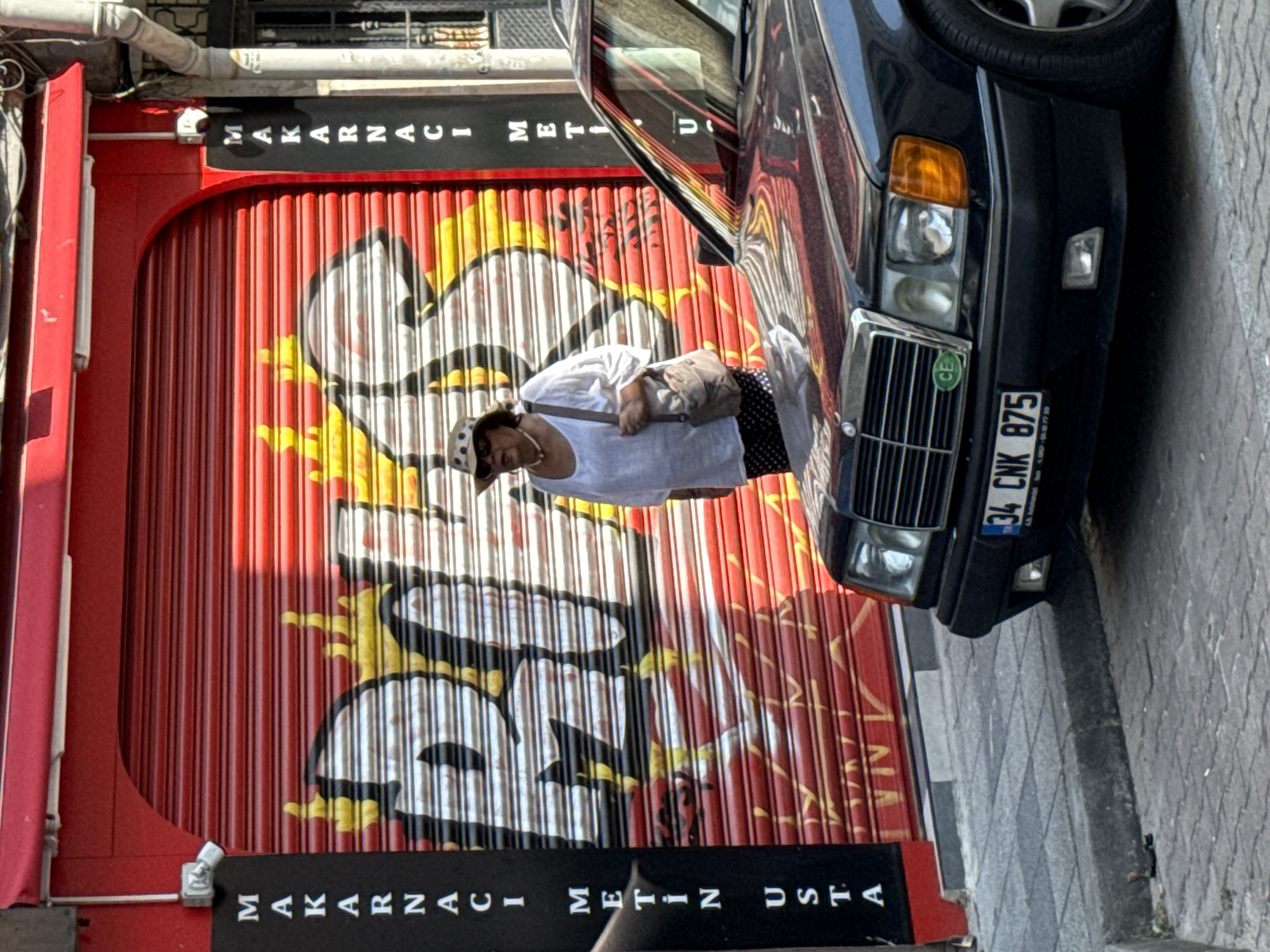 A woman wearing a patterned hat, sunglasses, a white tunic, and dark polka-dot pants stands in front of a red metal security shutter with graffiti of yellow flames and white lettering that appears to read "BULGUR". She is carrying a light brown shoulder bag. A dark blue Mercedes-Benz sedan is parked in front of the shutter, its front end visible. The car has a license plate that reads "34 CNK 875". A black vertical sign with white lettering that reads "MAKARNACIMETIN USTA" is on the left side of the shutter. The scene is on a paved sidewalk with a gray curb in front of the shutter. The location is Kadıköy, Turkey.