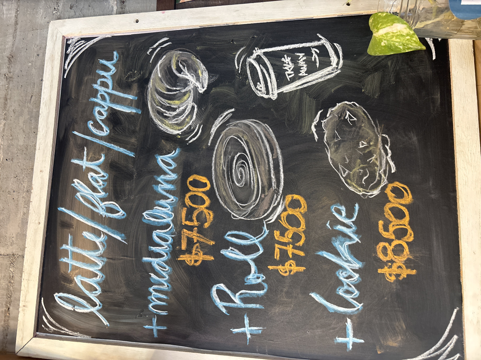 A close-up shot of a chalkboard menu with a white wooden frame, the background is a dark slate. The menu items, drawn in white chalk, include a pastry resembling a croissant ("medialuna"), a spiral roll, and a cookie, each accompanied by a corresponding illustration in white chalk. The prices for each item are written in orange chalk and appear to be "$7500" for the medialuna and roll, and "$8500" for the cookie. The words "latte/flat/cappu" are written vertically along the left side in a light blue chalk, suggesting coffee offerings. The words "TAKE AWAY" are included in the drawing. The chalkboard is angled slightly towards the upper right. A green leaf is visible at the top right of the frame. The menu implies a café setting, likely in Buenos Aires, Argentina, based on the common availability of medialunas.