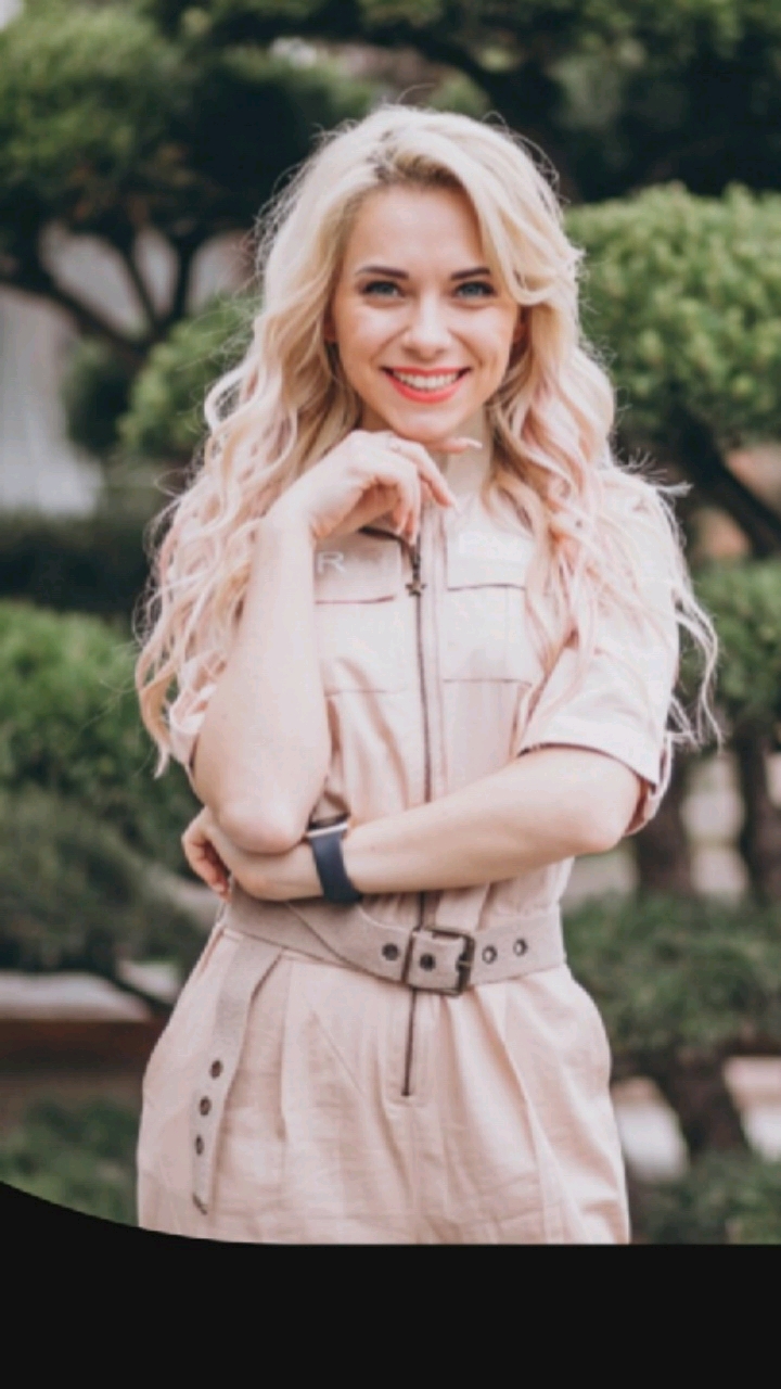 A young woman with blonde, wavy hair and blue eyes smiles directly at the camera. She is wearing a light pink, collared jumpsuit with a belt around her waist. Her arms are crossed, and her left hand rests on her chin. The background consists of blurred green trees and foliage, suggesting an outdoor setting. The image is a medium shot, focusing on the woman's upper body and face. There is no indication of the city of Hongkou District, China in the image.