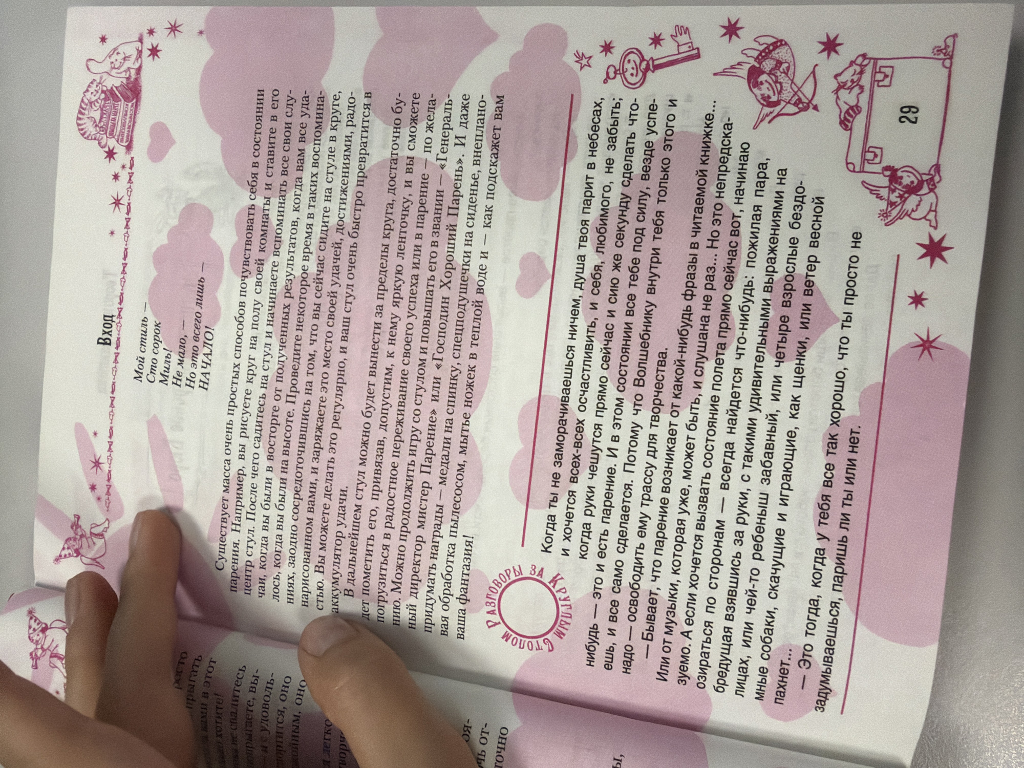 The image shows an open book with pink pages and illustrations. The book is likely a children's book or a book about imagination and creativity, given the whimsical drawings and playful text.
The left page contains handwritten text in black marker, partially obscuring the printed text. The printed text is in Russian. The right page also has printed text in Russian and several illustrations.
Illustrations include:
* Pink clouds and stars scattered across both pages.
* A decorative border with winged cherubs holding bows and arrows.
* A cat sleeping on a stack of books and a rabbit-like creature on the right page.
* A circular stamp with the words "РАЗГОВОРЫ ЗА КРУГОМ" (Conversations around the circle) on the left page.
* A drawing of a key with wings and a decorative element on the right page.
* A suitcase with the number "29" on it, with a winged creature and a cat on top, also on the right page.
The open book is lying flat, and a human hand is visible at the top left corner of the image, holding the book open. The lighting suggests an indoor environment, possibly a room. The overall aesthetic is lighthearted and imaginative.
There is no information in the image that provides any details about the location in the city of Homyel, Belarus.