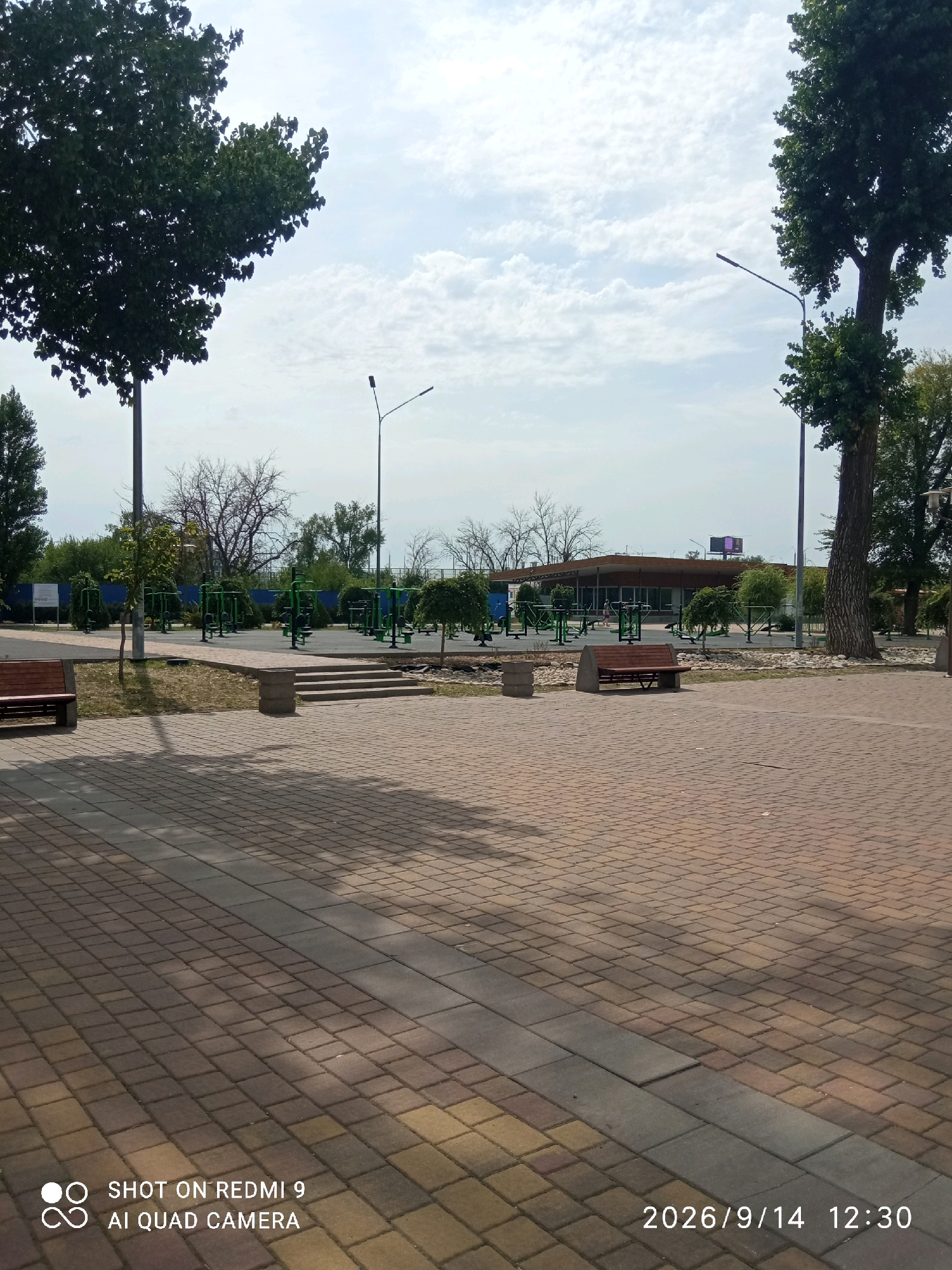 This image captures a daytime outdoor scene at a public recreational area in Rostov-on-Don, Russia, likely a park or promenade. The foreground is dominated by a paved area with interlocking brickwork in shades of brown and grey, casting long shadows from unseen objects.
In the middle ground, there is a dedicated outdoor fitness zone featuring multiple green metal exercise machines arranged in a structured layout on a grey paved surface. A set of concrete stairs leads up to this area from the brick-paved section. Two wooden benches with metal frames are visible, one near the stairs and another further to the right. Several young trees and bushes are planted around the fitness equipment and pathways.
In the background, a long, low-rise building with a flat roof and large windows is situated. A large digital display board with a purple graphic is mounted on the building. Beyond the building, trees and vegetation form a natural backdrop. Two tall lampposts with single lights are also present, one on the left and one on the right side of the image.
The sky is partly cloudy with scattered white clouds against a pale blue backdrop. Two large, mature trees frame the scene on the left and right sides, their dense green foliage contrasting with the sky. The overall impression is of a well-maintained public space designed for recreation and exercise. The timestamp in the lower right corner indicates "2026/9/14 12:30".