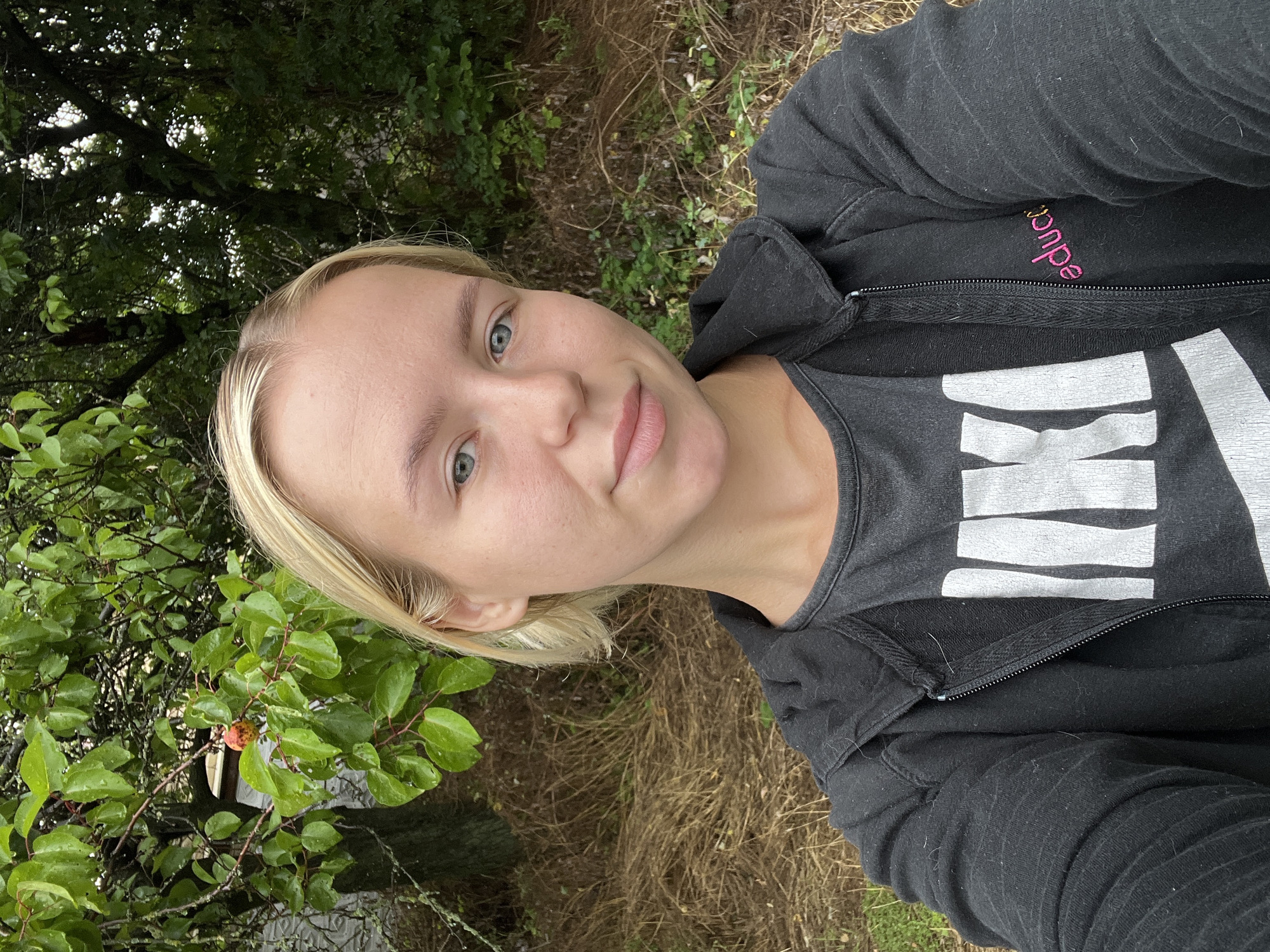 A young, fair-skinned woman with short blonde hair takes a selfie outdoors. She has blue eyes and light-colored eyebrows, and she is not wearing makeup. She is wearing a dark gray t-shirt with a white logo that appears to be Nike, and a black zip-up hoodie over it. The hoodie has a small pink embroidered logo on the right side. Behind her, a tree with green leaves and a single reddish-orange fruit is visible, along with dry, brown grass and other greenery. The lighting suggests it is daytime, possibly overcast.