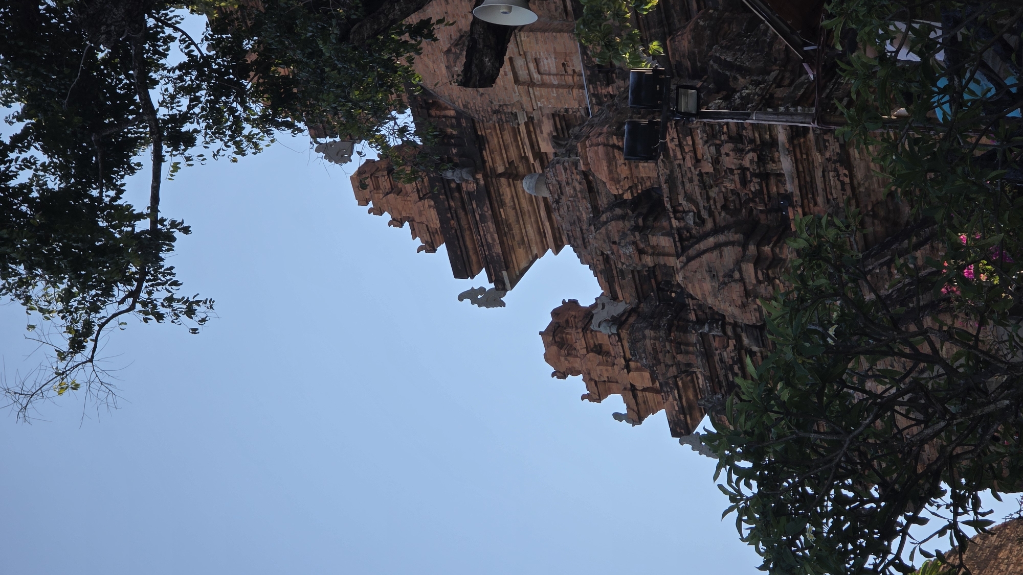 A ground-level, upward-facing shot captures a portion of the Po Nagar temple complex in Bắc Nha Trang, Vietnam. The camera angle gives prominence to the red brick structures of the temple. The clear blue sky forms a backdrop for the angular architecture. Trees are seen at the top left and right of the image, framing the temple structure. A modern light fixture is attached to the temple wall, suggesting the site is still in use.