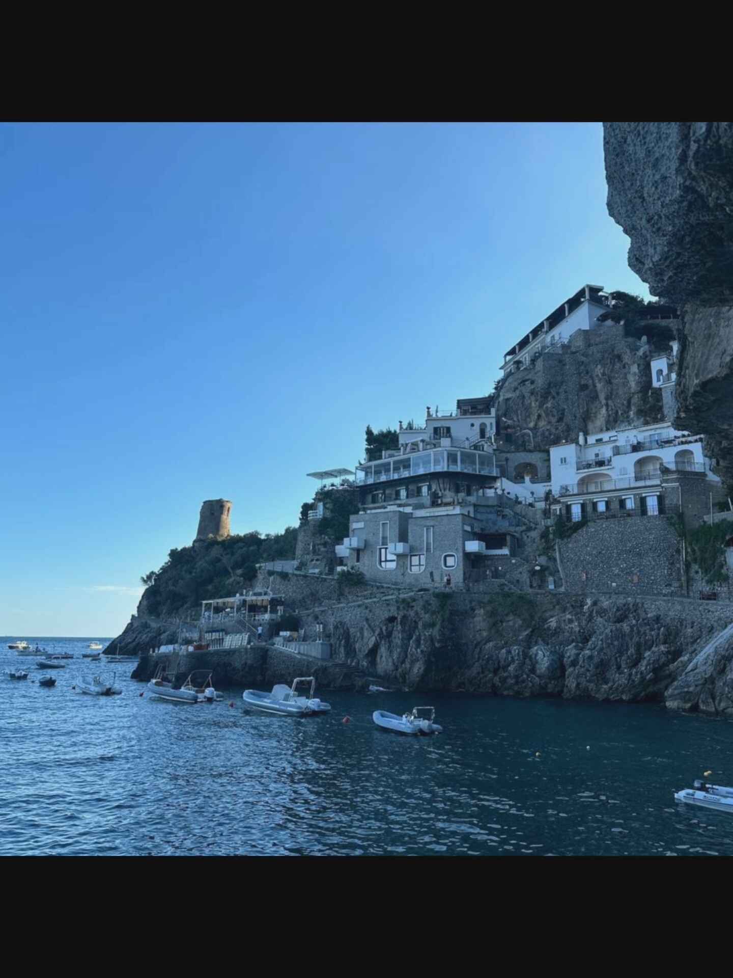 The image depicts a coastal scene with buildings clinging to a steep cliffside, overlooking a body of water. The architecture is characterized by white, multi-story structures with balconies and large windows, integrated into the rocky terrain. Several boats, including what appear to be rigid inflatable boats (RIBS), are moored in the water. A prominent stone tower stands on a higher elevation of the cliff. The sky is a clear, bright blue. The scene suggests a Mediterranean coastal town. The prompt mentions the city of Norcia, Italy, however, Norcia is an inland town and does not have a coastline. This scene is likely not in Norcia, Italy.