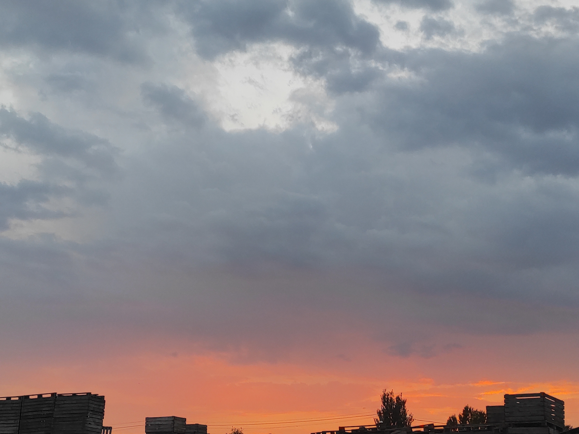 The image shows a twilight sky over an industrial or agricultural area. The sky is a gradient of colors, transitioning from dark gray clouds at the top to a vibrant orange and pink sunset at the horizon. Silhouetted against the colorful sky are rows of wooden crates or structures, some appearing to be stacked high. Trees and power lines are also visible along the horizon. The overall scene suggests a rural or semi-industrial setting at dusk.